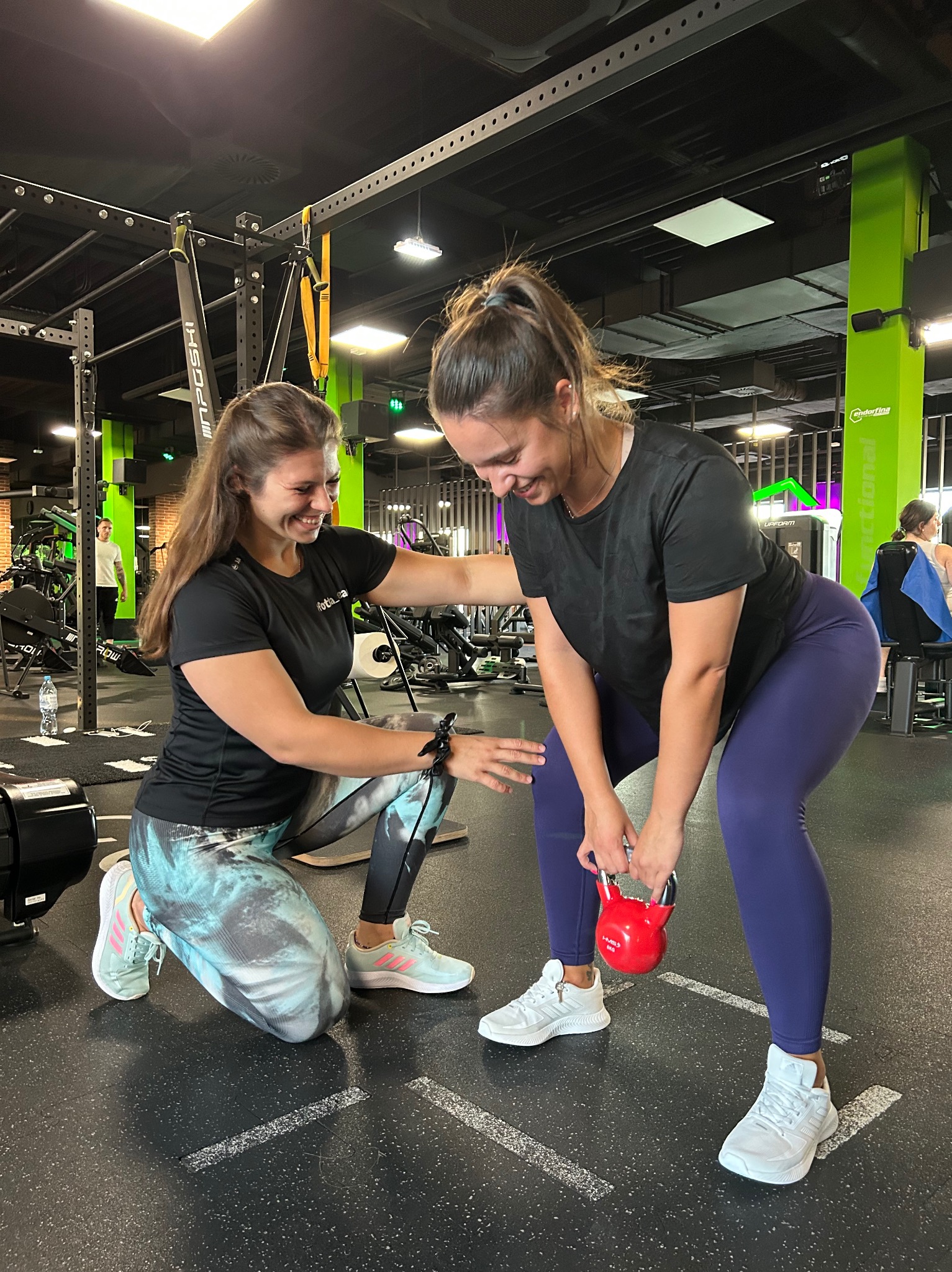 Trenerka personalna nadzoruje ćwiczenie z kettlebell, uśmiechnięta kobieta w sportowym stroju w trakcie treningu na tle siłowni.