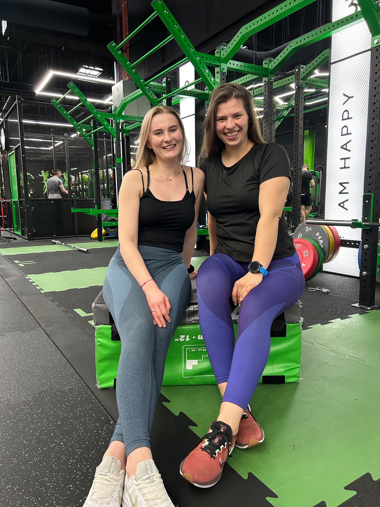 Dwie uśmiechnięte kobiety w sportowych strojach siedzą na skrzyni w nowoczesnej siłowni z zielonymi elementami konstrukcyjnymi i napisem 'I AM HAPPY'.