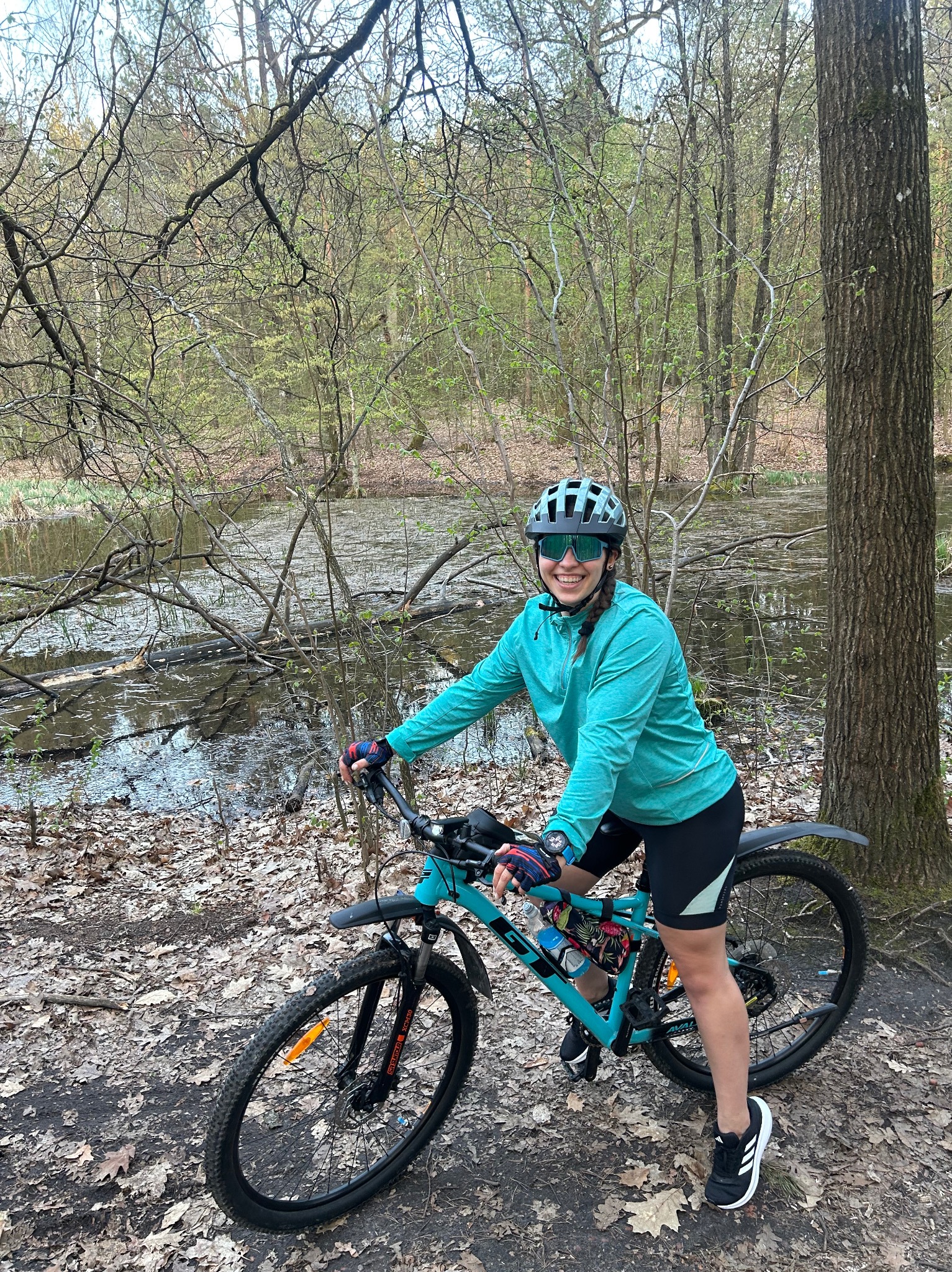 Uśmiechnięta kobieta w kasku i okularach na rowerze górskim, w tle leśne jeziorko, na ziemi liście. Aktywny wypoczynek na łonie natury.