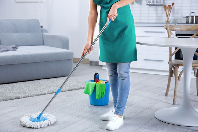 Osoba w zielonym fartuchu myje podłogę mopem w jasnym pokoju z szarą kanapą i okrągłym stołem, wiadro ze środkami czystości obok.