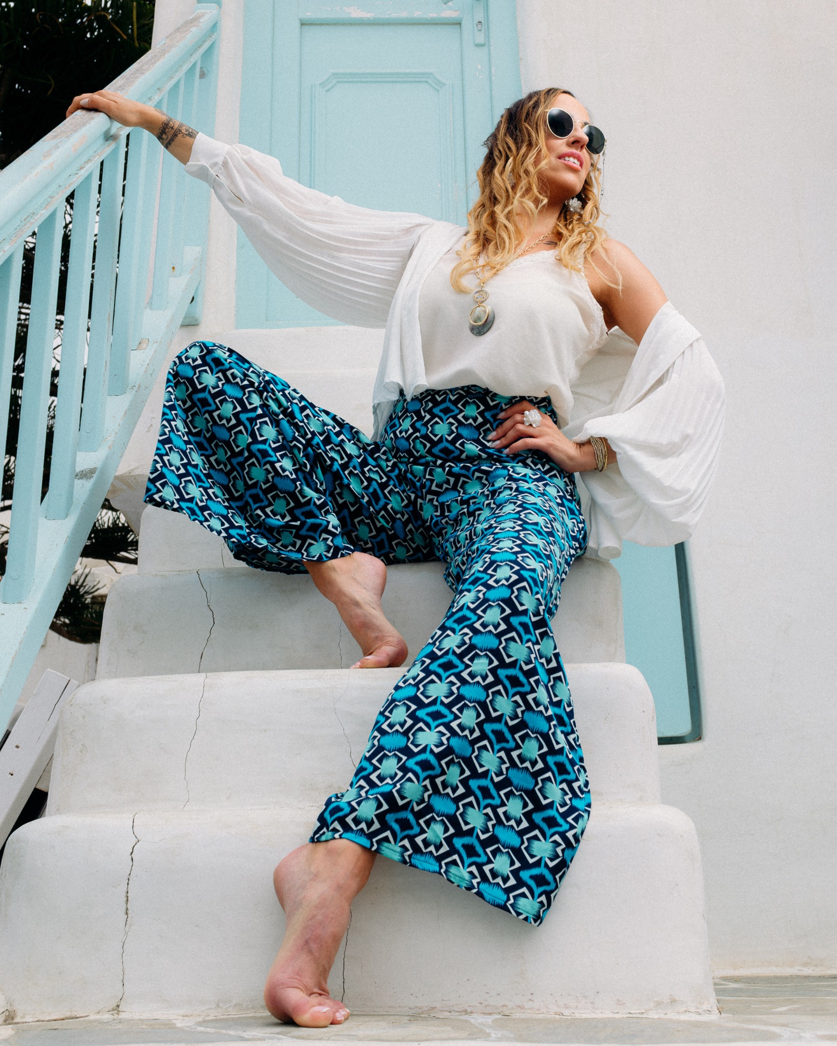Kobieta w okularach przeciwsłonecznych, białej bluzce z bufiastymi rękawami i wzorzystych, szerokich spodniach siedzi na białych schodach z niebieską barierką i drzwiami w tle, pozując z uniesioną...
