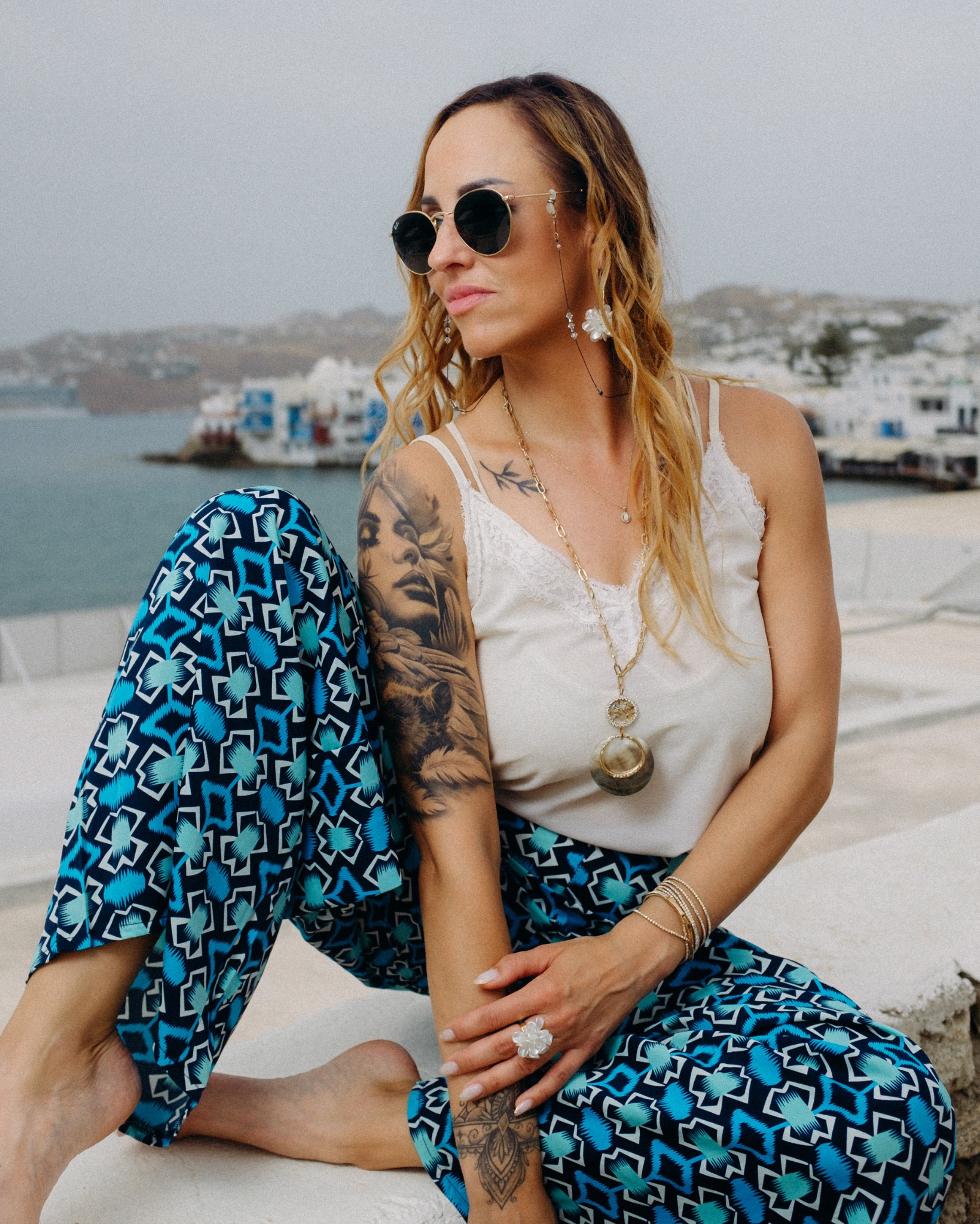 Kobieta w okularach przeciwsłonecznych siedzi na tle morza i białych budynków, ubrana w biały top i wzorzyste niebieskie spodnie, z widocznymi tatuażami na ramieniu i stopie oraz biżuterią.