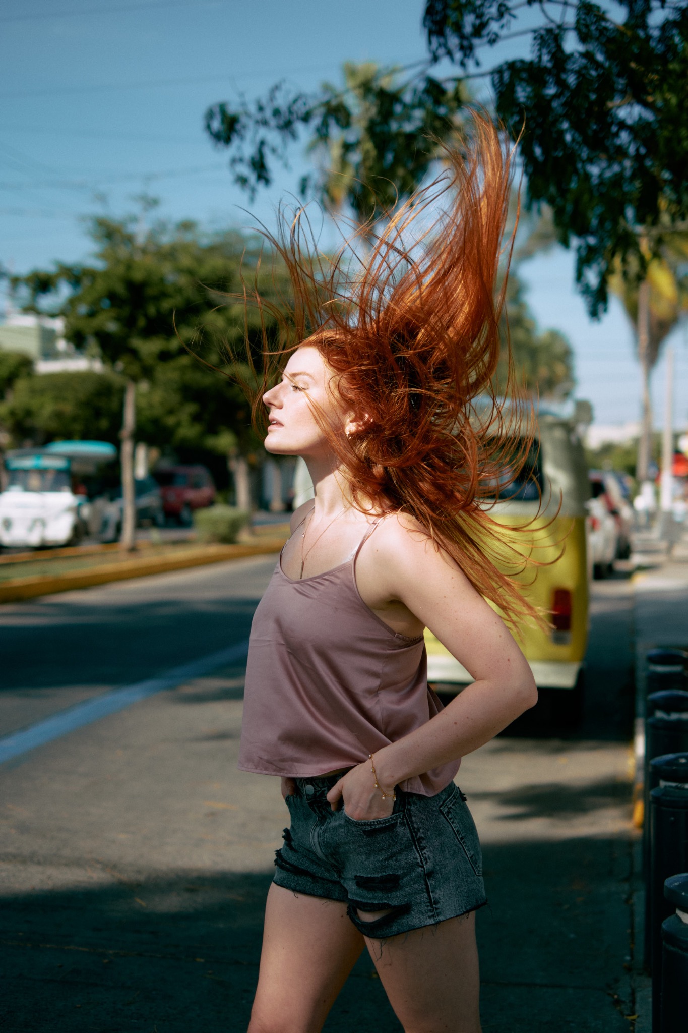 Ruda kobieta w szortach i topie odchyla głowę do tyłu, a jej włosy unoszą się w powietrzu na tle ulicy z samochodami i zielenią.