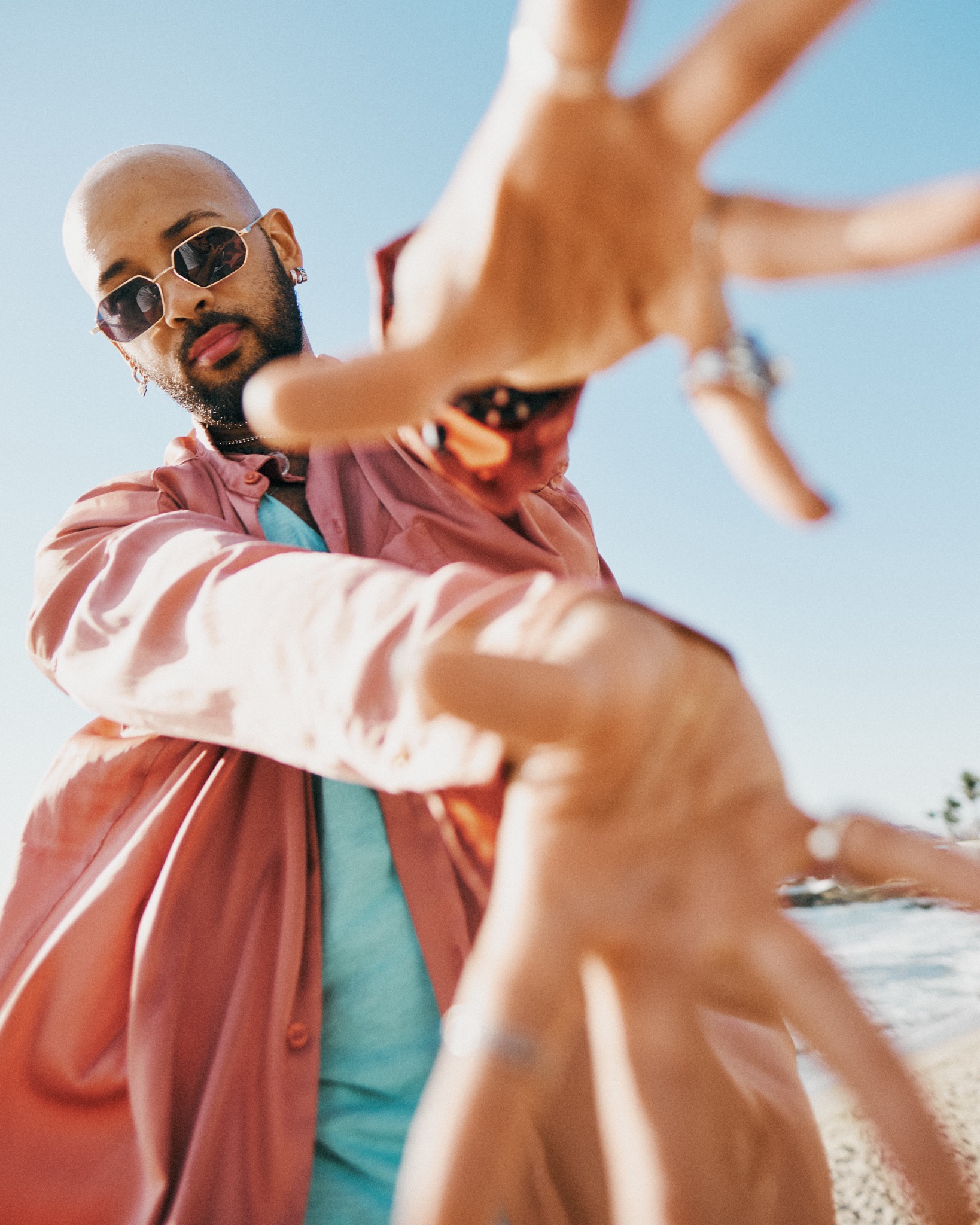 Mężczyzna w okularach i różowej koszuli pozuje na plaży, wyciągając dłonie w stronę obiektywu.