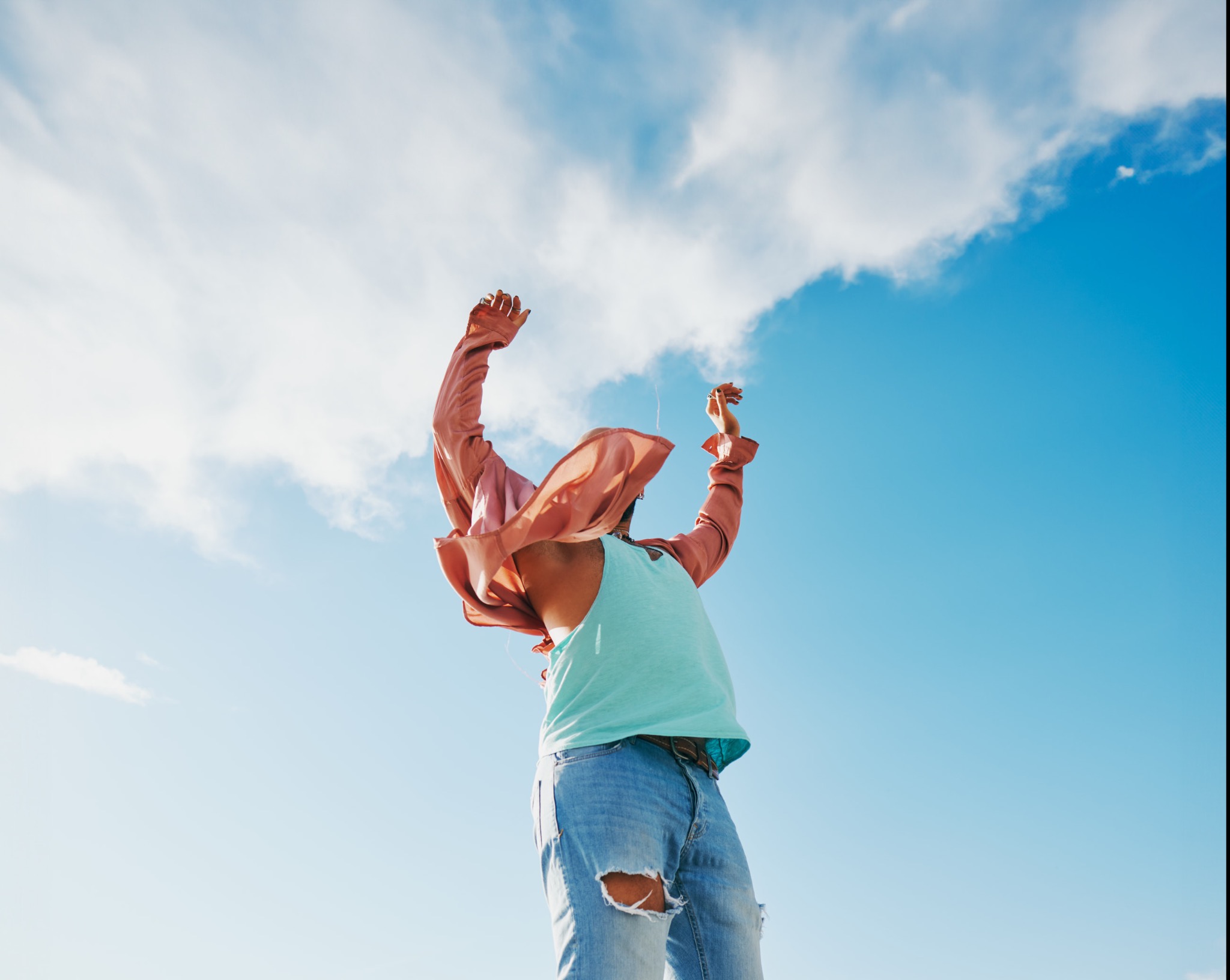 Osoba w dżinsach z dziurami i różowej koszuli, z rękami uniesionymi do góry na tle błękitnego nieba z chmurami