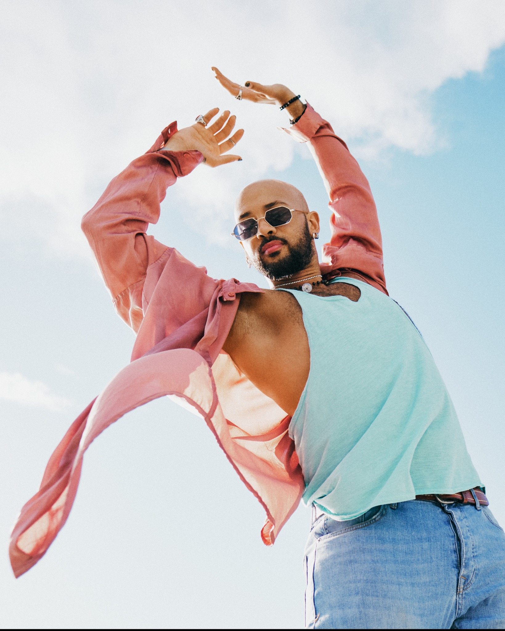 Mężczyzna w okularach przeciwsłonecznych, różowej koszuli i błękitnym podkoszulku, pozujący na tle jasnego nieba.