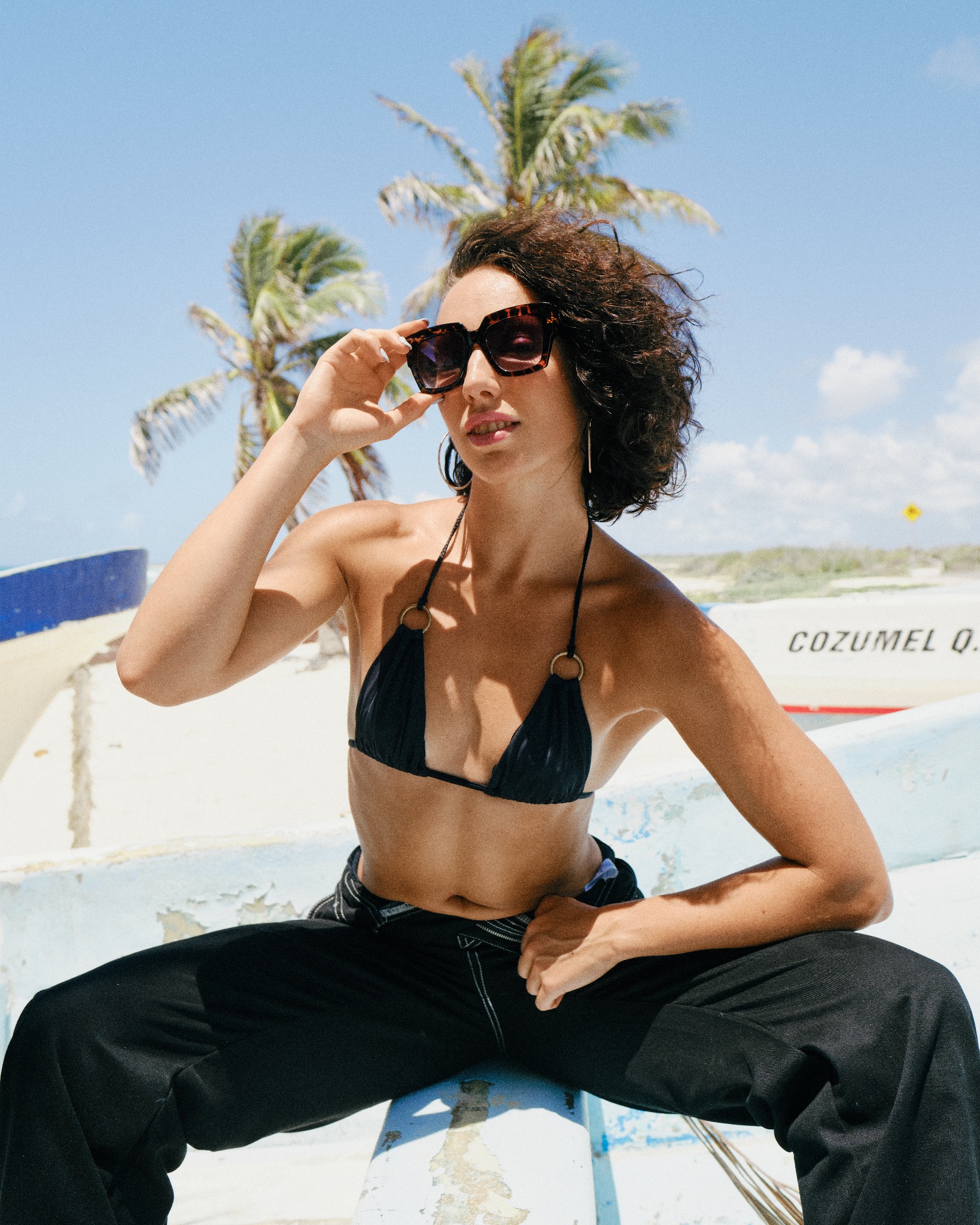 Kobieta w bikini i spodniach siedząca na łodzi z napisem Cozumel Q., w tle palmy i błękitne niebo.