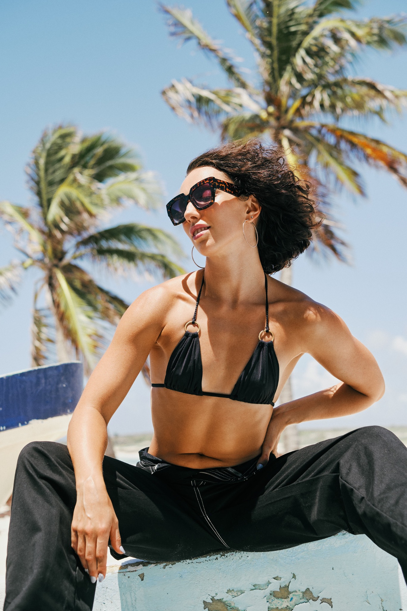 Kobieta w czarnym bikini i spodniach siedzi na niebieskiej betonowej powierzchni, z palmami w tle, spoglądając w lewo.