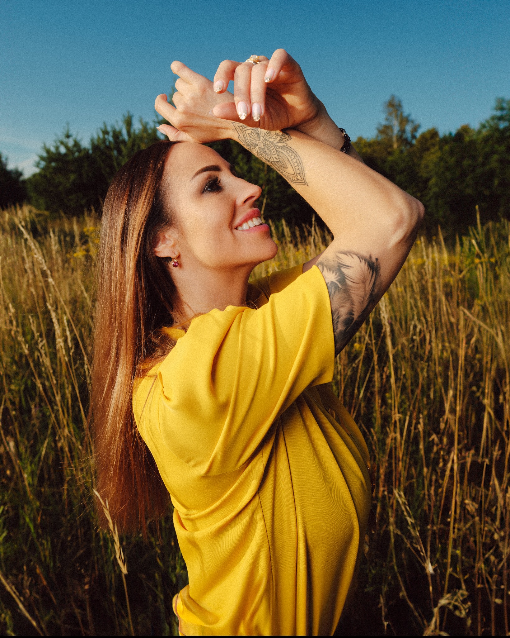 Portret uśmiechniętej kobiety w żółtej sukience, stojącej na polu w słoneczny dzień, z rękami uniesionymi nad głową, eksponującymi tatuaże na ramionach i dłoniach, oraz pierścionek na palcu.