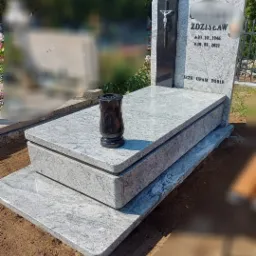 Jasnoszary, dwupoziomowy pomnik z granitu z czarnym krzyżem i wazonem, inskrypcjami oraz wygładzonymi krawędziami, otoczony ziemią i zielenią cmentarza.