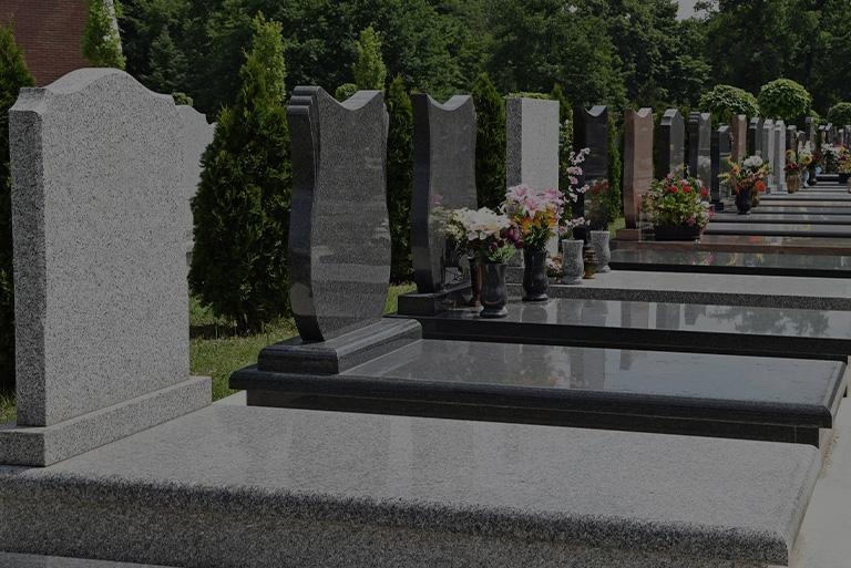 Rząd granitowych pomników z kwiatami w wazonach, ustawionych na cmentarzu, z zielenią drzew w tle.