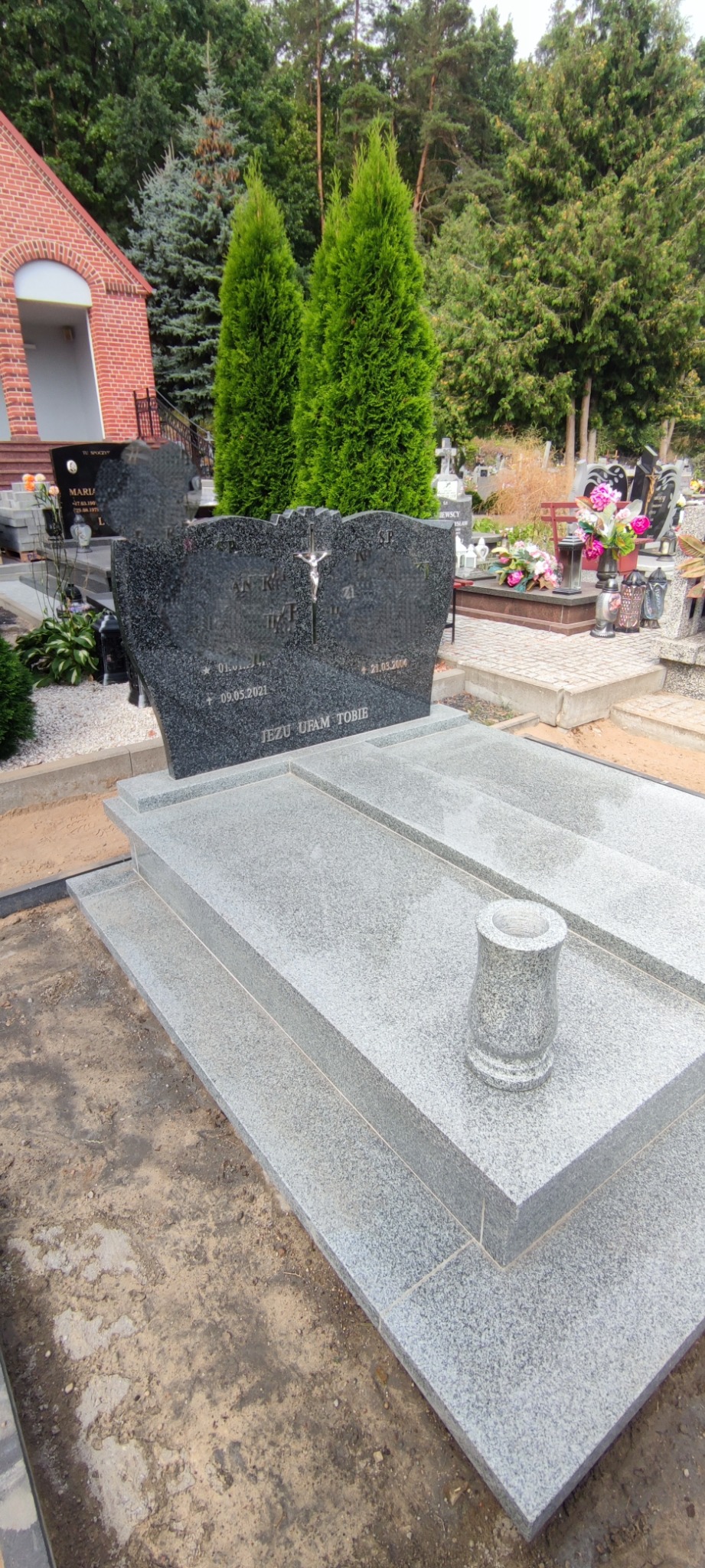 Granitowy nagrobek z ciemną tablicą inskrypcyjną i jasnoszarą podstawą, w tle inne nagrobki i zieleń cmentarza.