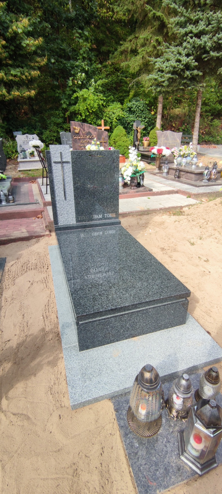 Nowoczesny, ciemnoszary pomnik z krzyżem, otoczony piaskiem, z widocznymi lampionami i nagrobkami w tle, na cmentarzu.