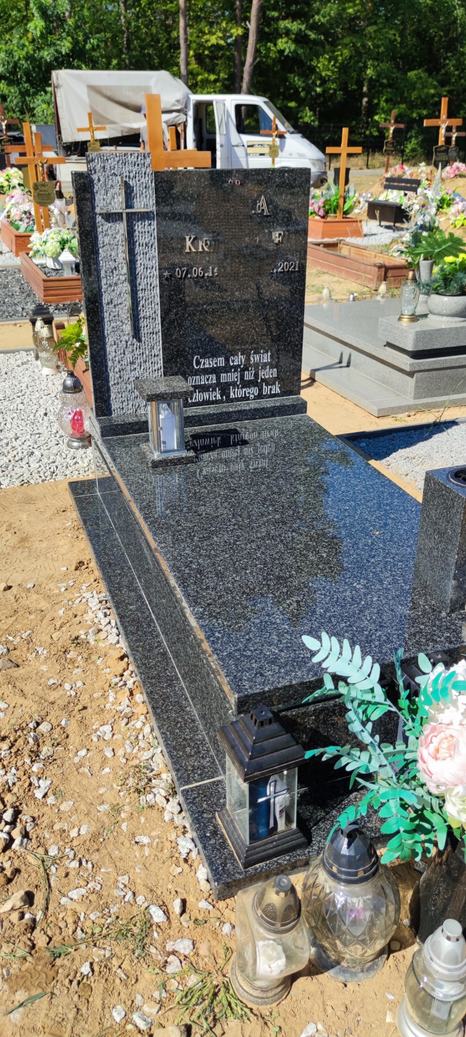 Czarny, polerowany pomnik z granitu na cmentarzu, z wygrawerowanym napisem i datami, ozdobiony lampionami i kwiatami, otoczony piaskiem i innymi grobami.