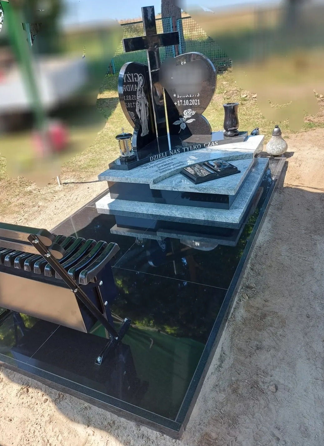 Czarny nagrobek z krzyżem i sercem, z wygrawerowaną postacią kobiety i datą, uzupełniony o metalową ławkę na czarnym, błyszczącym fundamencie.