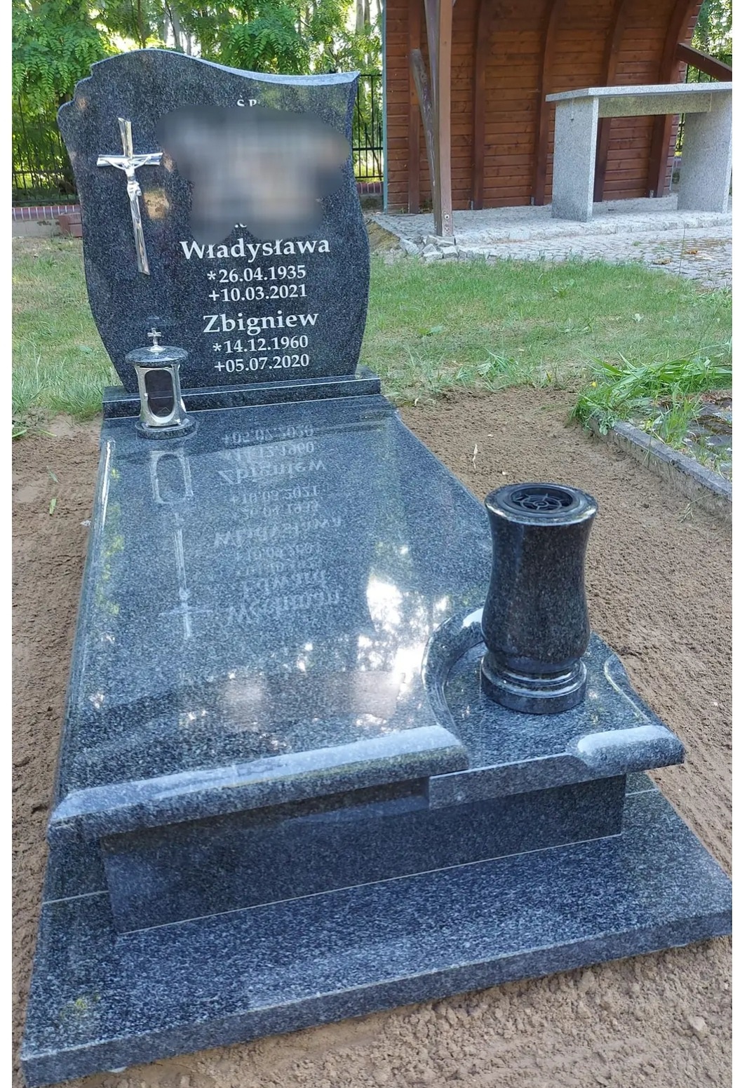 Granitowy pomnik z krzyżem i lampionem, widoczne daty urodzin i śmierci, w tle fragment drewnianej konstrukcji.