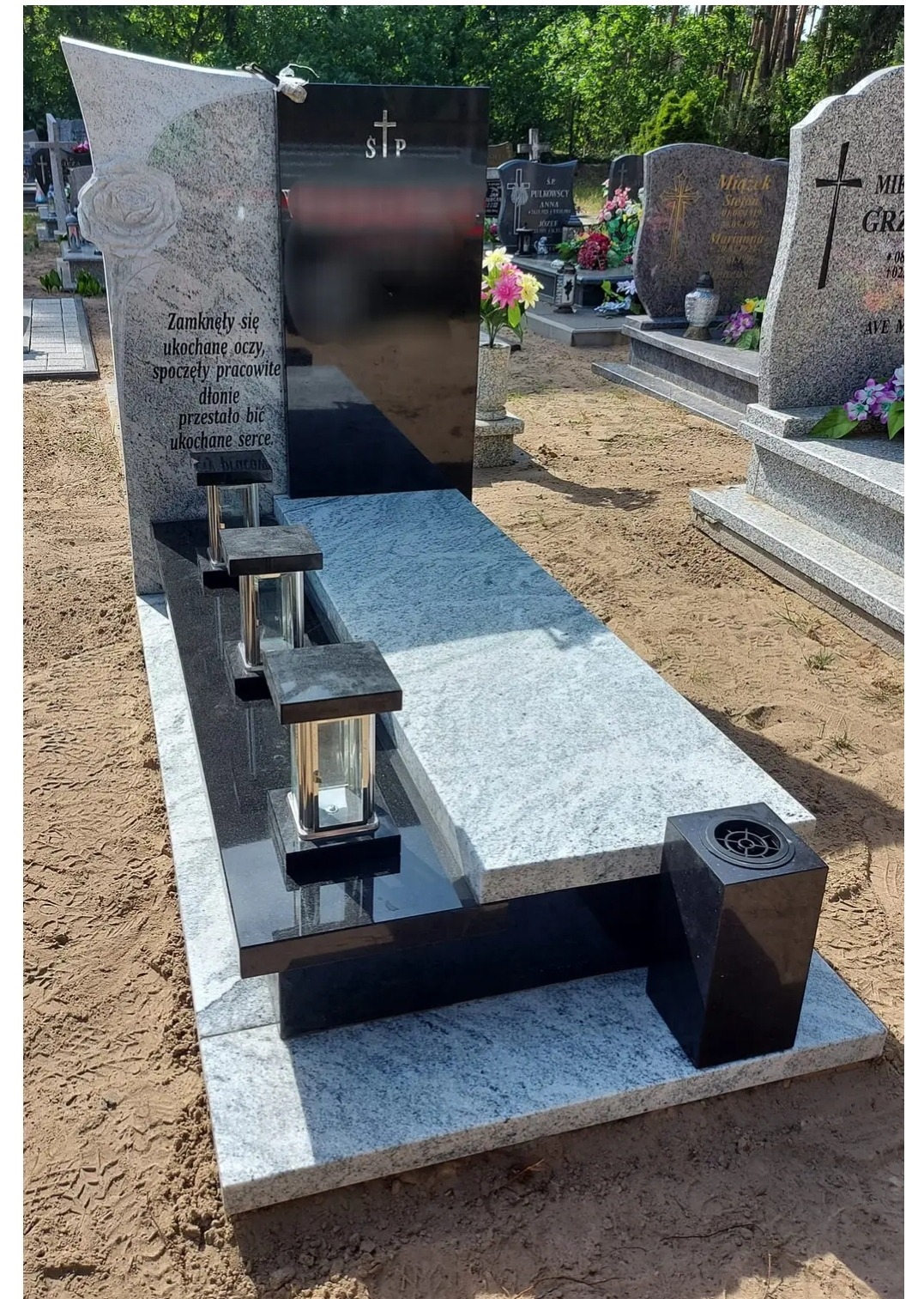Nowoczesny, dwukolorowy pomnik z jasnoszarego i czarnego granitu na cmentarzu, z wygrawerowaną różą i sentencją, z trzema lampionami w metalowych obudowach i czarnym pojemnikiem na kwiaty.