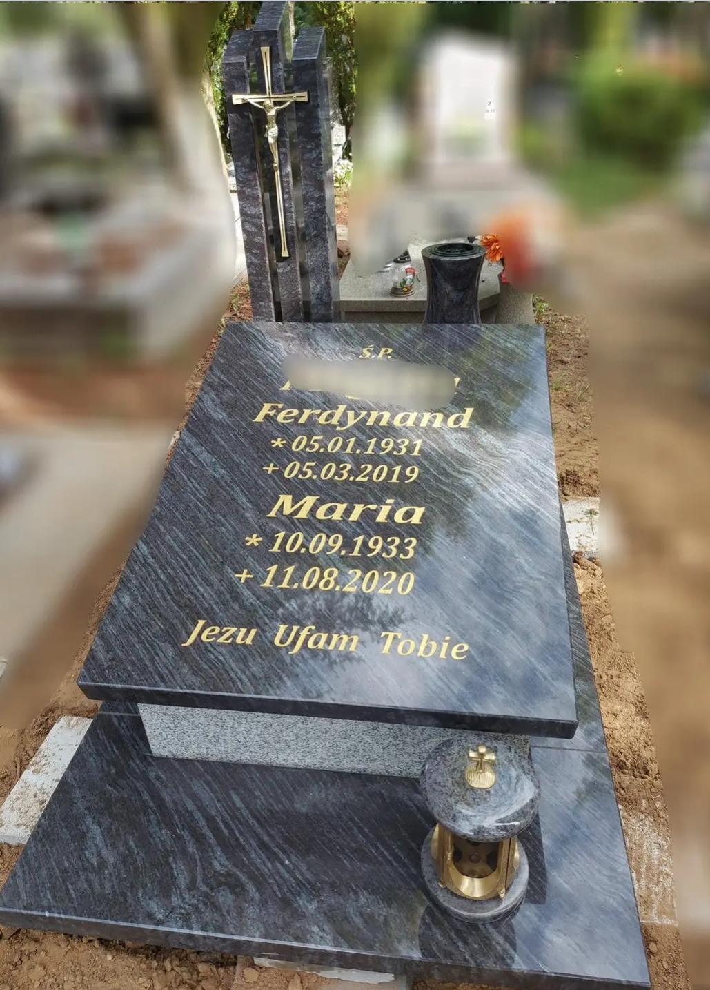 Granitowy pomnik z wygrawerowanymi złotymi literami imion i dat z krzyżem w tle, ustawiony na cmentarzu.