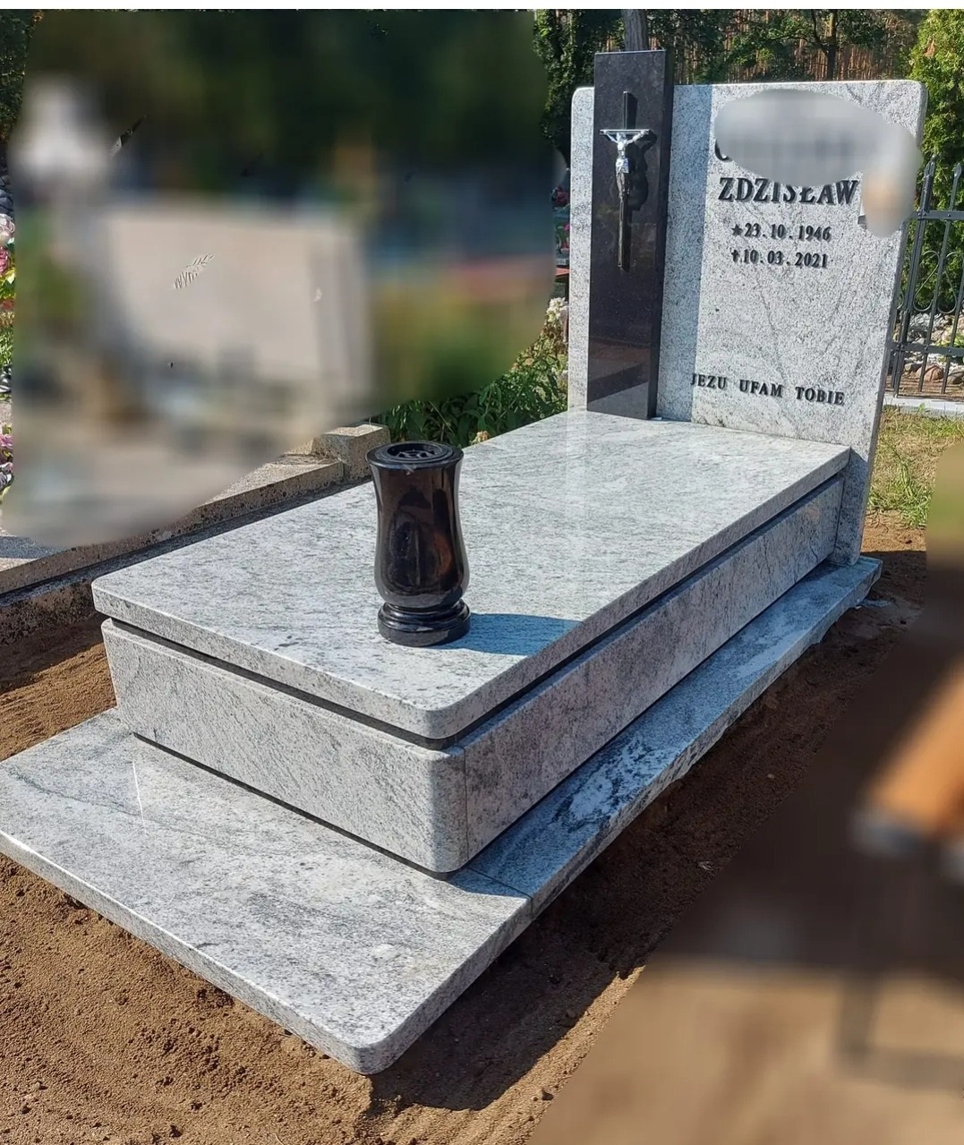 Jasnoszary, dwupoziomowy pomnik z granitu z czarnym krzyżem i wazonem, inskrypcjami oraz wygładzonymi krawędziami, otoczony ziemią i zielenią cmentarza.