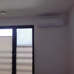 Biały klimatyzator zamontowany na ścianie nad oknem z żaluzjami plisowanymi w nowoczesnym wnętrzu mieszkalnym.