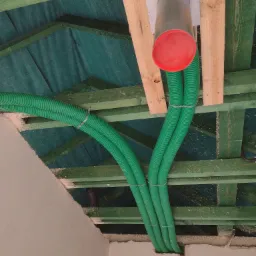 Zielone, karbowane rury wentylacyjne prowadzone pod stropem z drewnianych belek, widoczny fragment instalacji z metalową puszką i czerwonym zakończeniem, surowy stan wykończenia wnętrza.