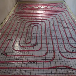 Czerwone rury ogrzewania podłogowego ułożone na folii z nadrukowaną siatką w pomieszczeniu z surowymi ścianami, widok z g&oacute;ry.