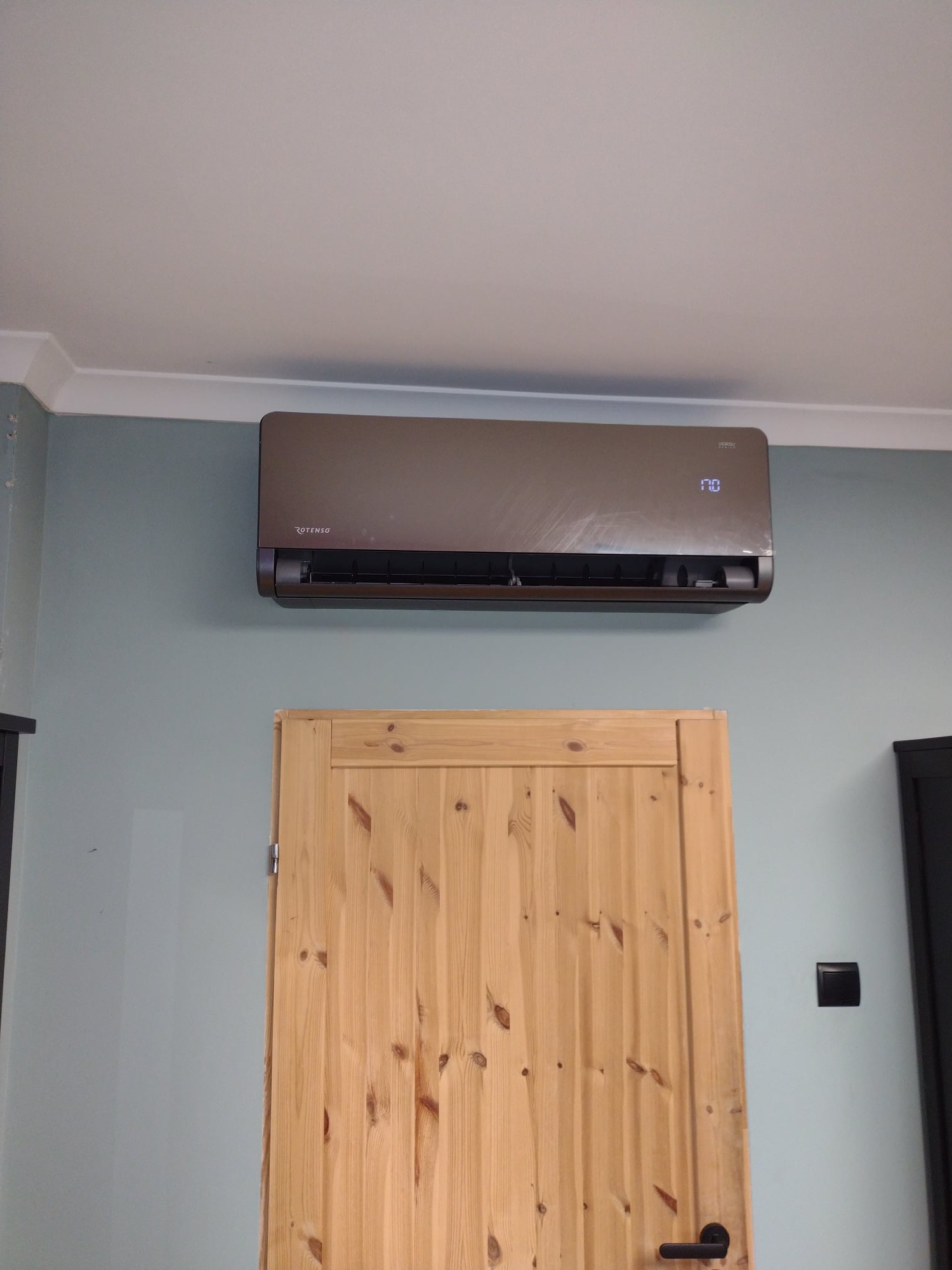 Brązowy klimatyzator ścienny Rotenso Versu z wyświetlaczem temperatury, zamontowany nad drewnianymi drzwiami w pomieszczeniu o jasnych ścianach.