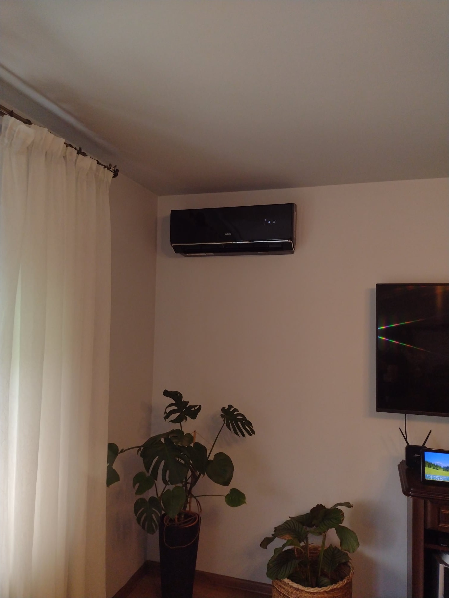Czarny klimatyzator ścienny marki AUX zamontowany w rogu pokoju, obok telewizora i roślin doniczkowych, podświetlony delikatnym światłem.