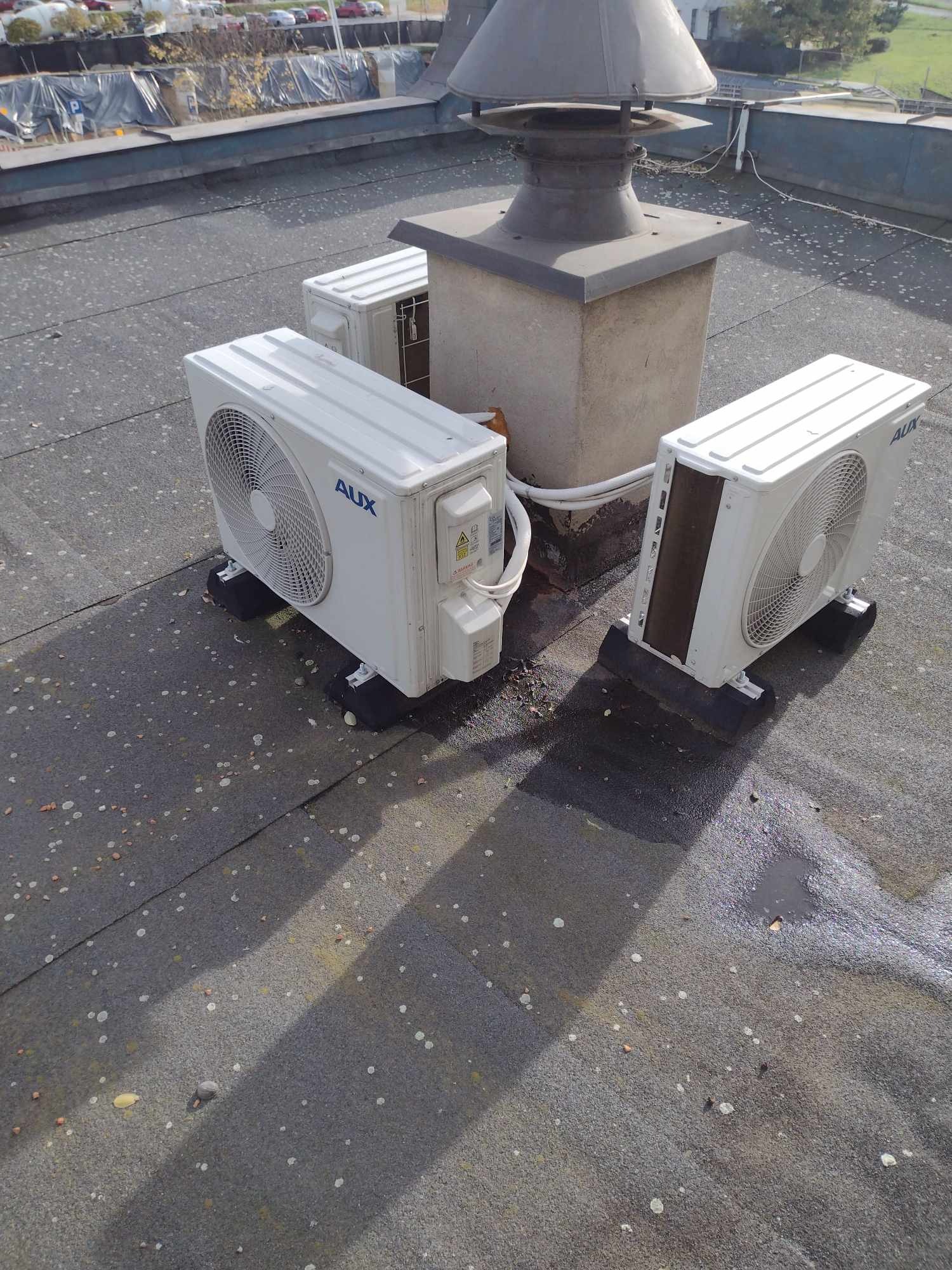 Trzy klimatyzatory marki AUX zainstalowane na płaskim, bitumicznym dachu obok komina wentylacyjnego.