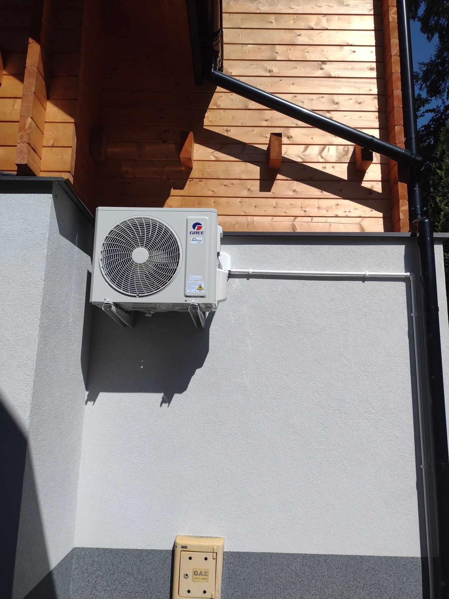 Zewnętrzna jednostka klimatyzacji marki Gree zamontowana na szarej ścianie budynku z drewnianą elewacją i czarną rynną.