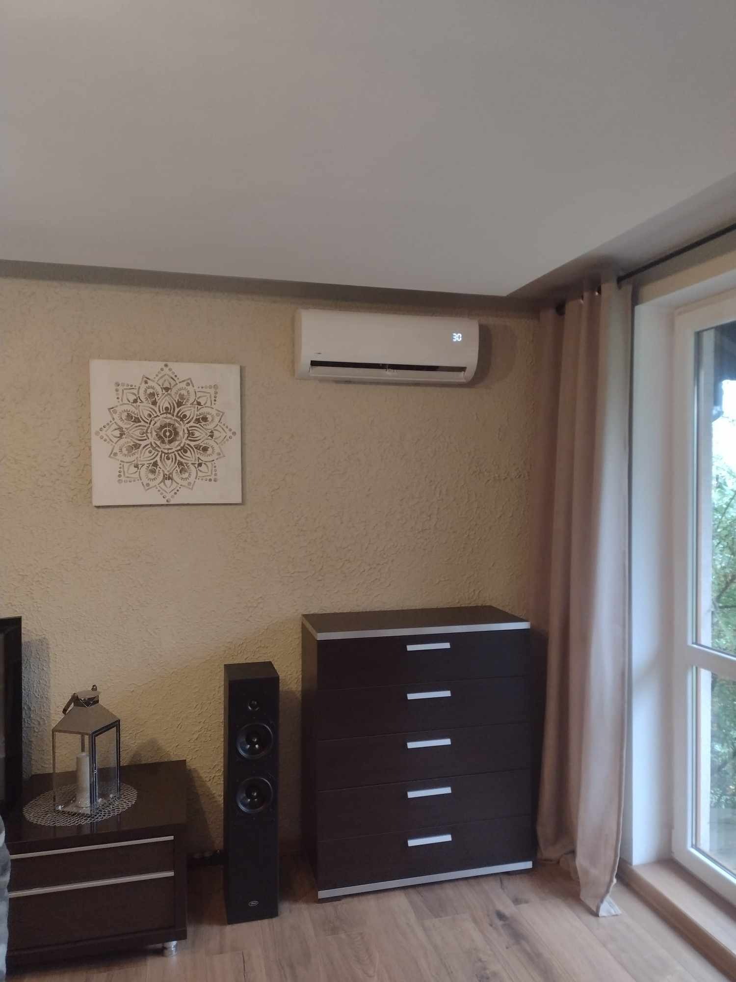 Wnętrze pokoju z zamontowanym klimatyzatorem ściennym, ustawionym na 30 stopni, obok obrazu z motywem mandali, z widoczną komodą, głośnikiem i stolikiem nocnym z dekoracyjną latarnią.