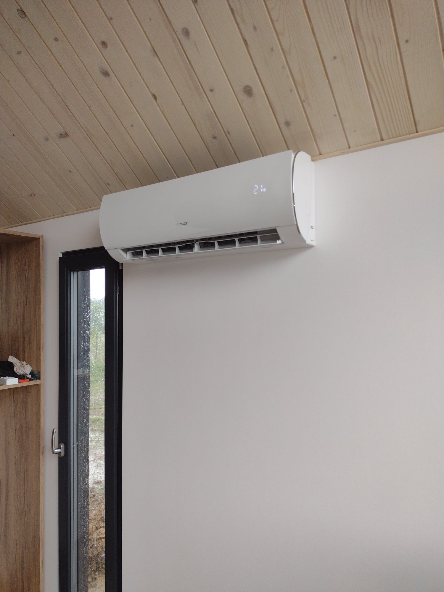 Biały klimatyzator ścienny z wyświetlaczem temperatury 21 stopni Celsjusza, zamontowany w pomieszczeniu z boazerią na suficie i oknem w czarnej ramie.