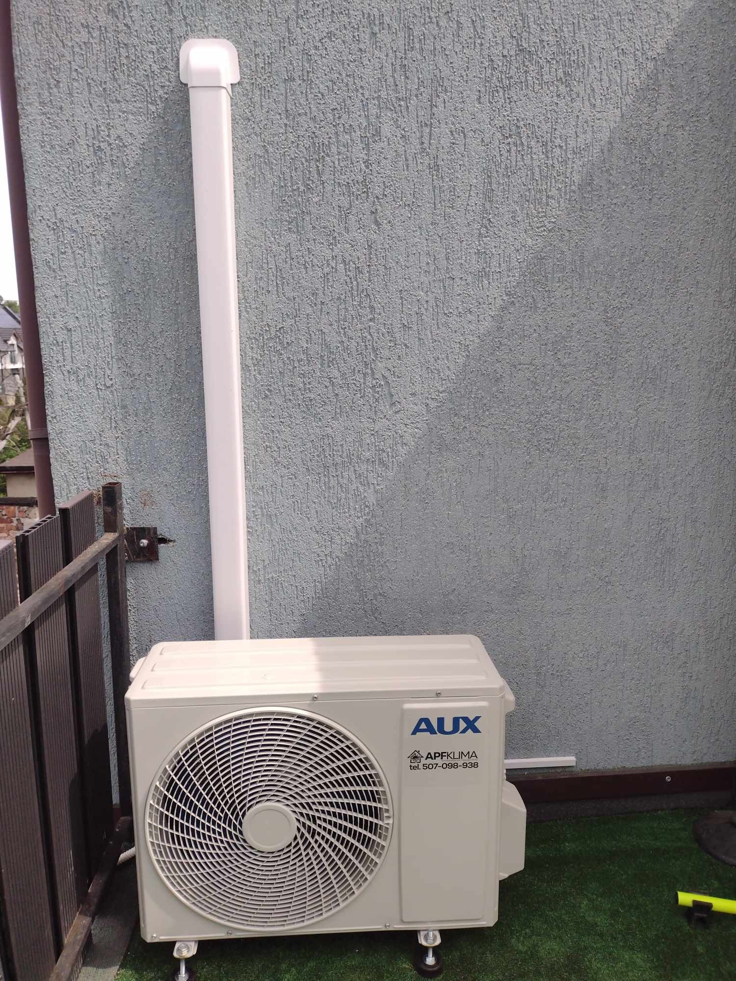 Zewnętrzna jednostka klimatyzacji AUX z odprowadzeniem skroplin po elewacji budynku o szarej, chropowatej fakturze, widoczna z boku na tle sztucznej trawy.