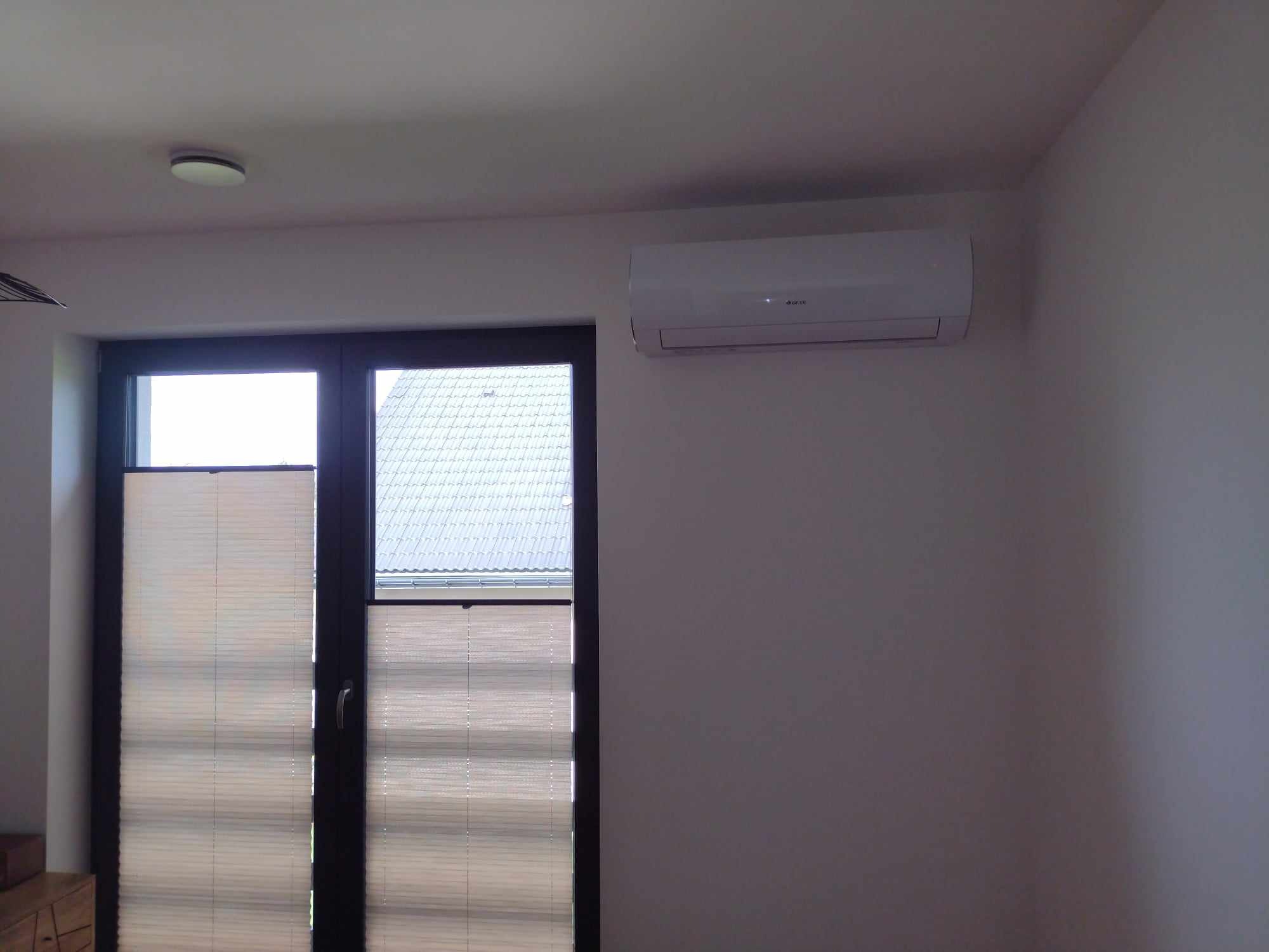 Biały klimatyzator zamontowany na ścianie nad oknem z żaluzjami plisowanymi w nowoczesnym wnętrzu mieszkalnym.