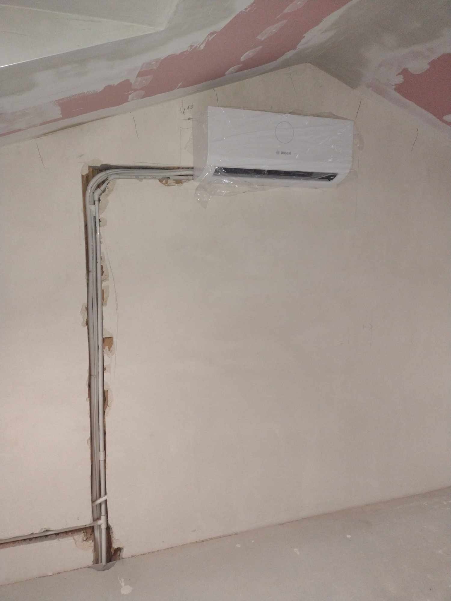 Nowy klimatyzator marki Bosch, jeszcze w folii ochronnej, zamontowany na ścianie pod skosem w surowym wnętrzu, widoczne rury instalacyjne w bruździe.