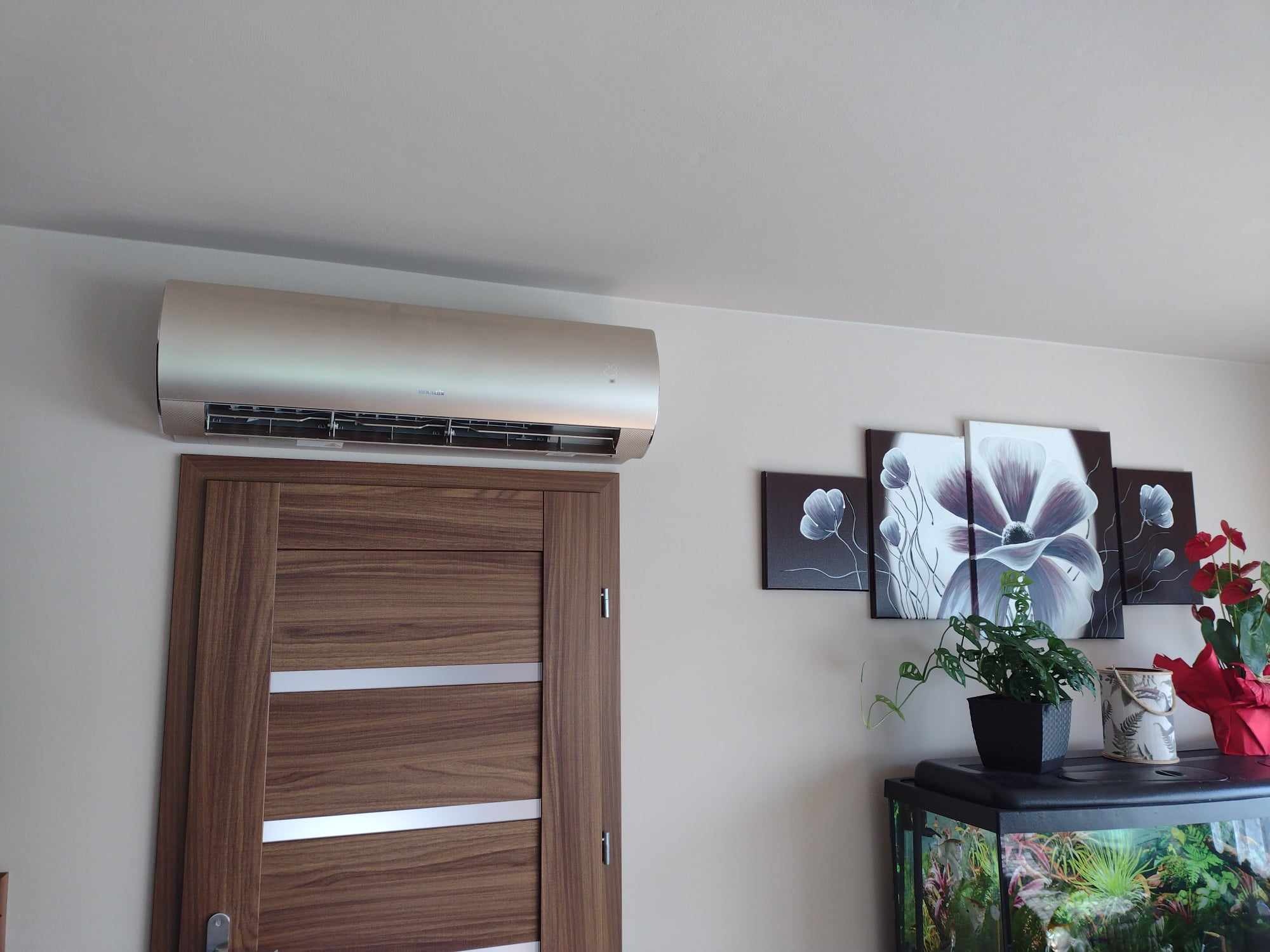 Nowoczesny klimatyzator w kolorze szampańskim zamontowany nad drewnianymi drzwiami z metalowymi wstawkami, obok obrazu z motywem kwiatowym i akwarium z roślinami.