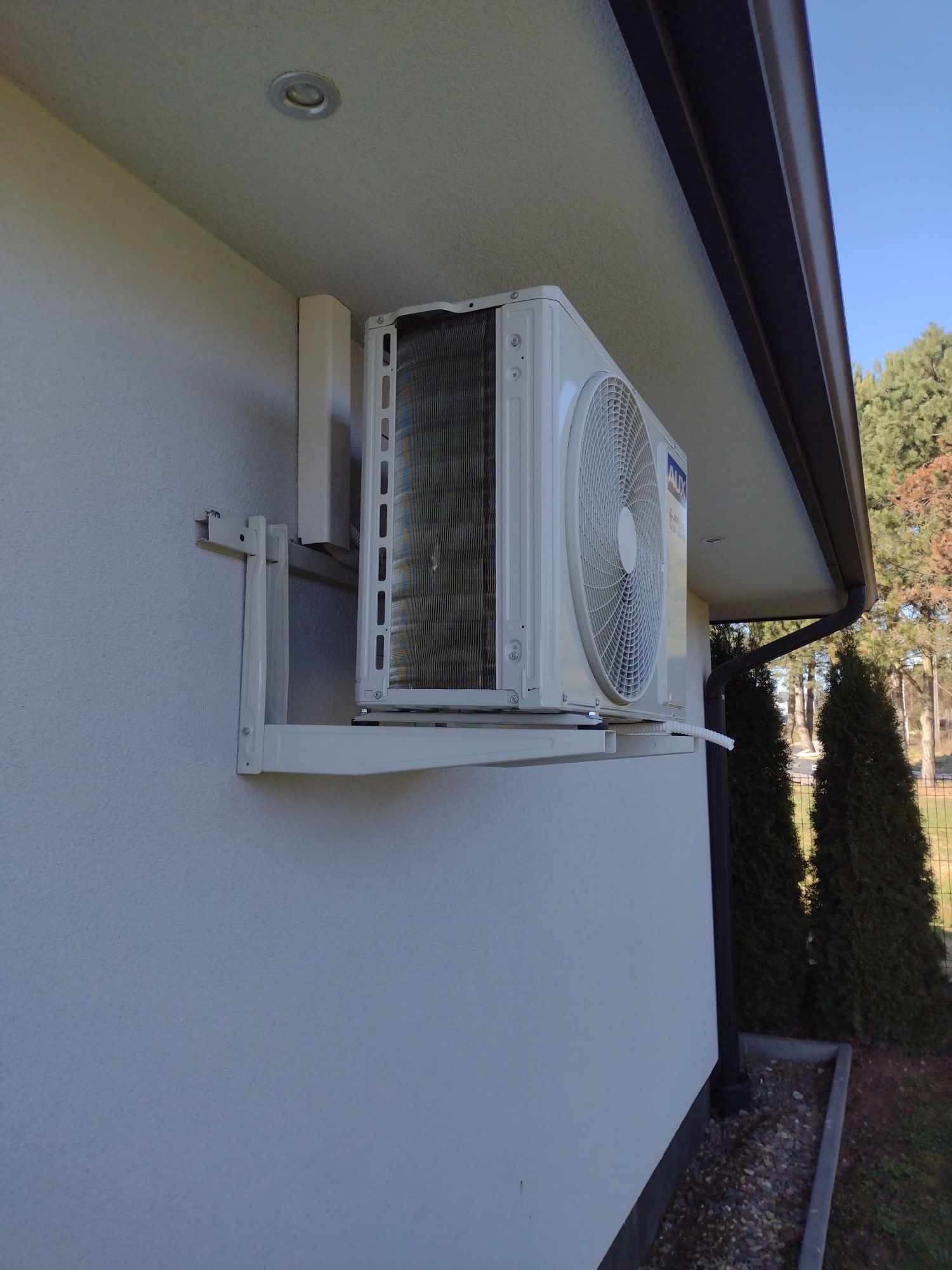 Zewnętrzna jednostka klimatyzacji marki AUX zamontowana na wsporniku na elewacji budynku jednorodzinnego, widoczne elementy instalacji i otoczenie z zielenią.