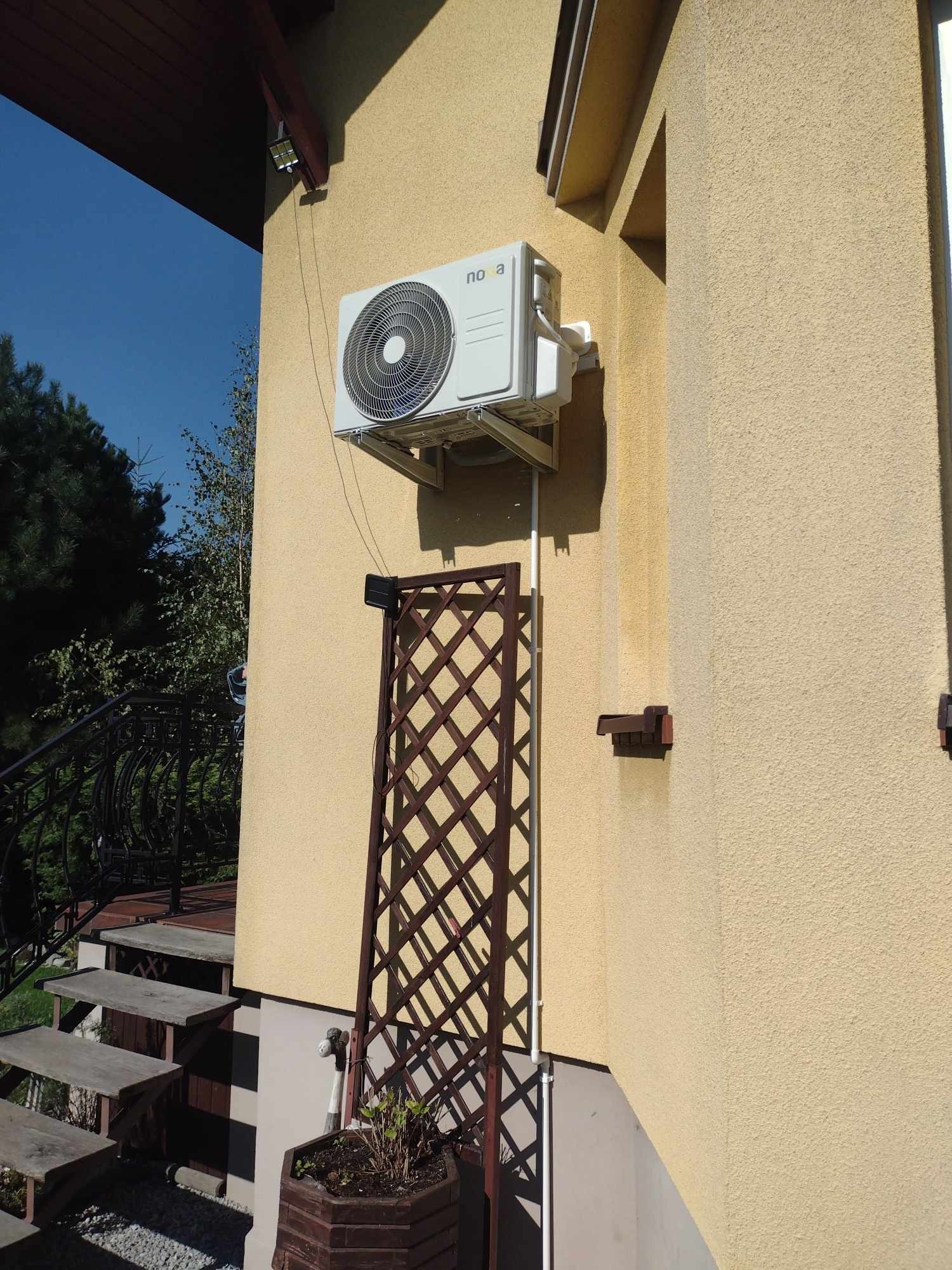 Biała jednostka zewnętrzna klimatyzatora marki Nova zamontowana na elewacji domu o żółtym odcieniu, widoczna krata dekoracyjna i schody wejściowe.