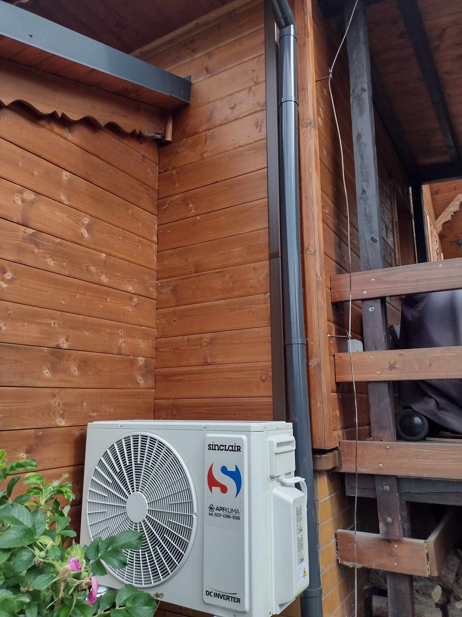 Zewnętrzna jednostka klimatyzacji Sinclair zamontowana na drewnianej ścianie domu, z widoczną rynną spustową i fragmentem drewnianej konstrukcji werandy oraz kwitnącym krzewem róży w dolnym rogu.