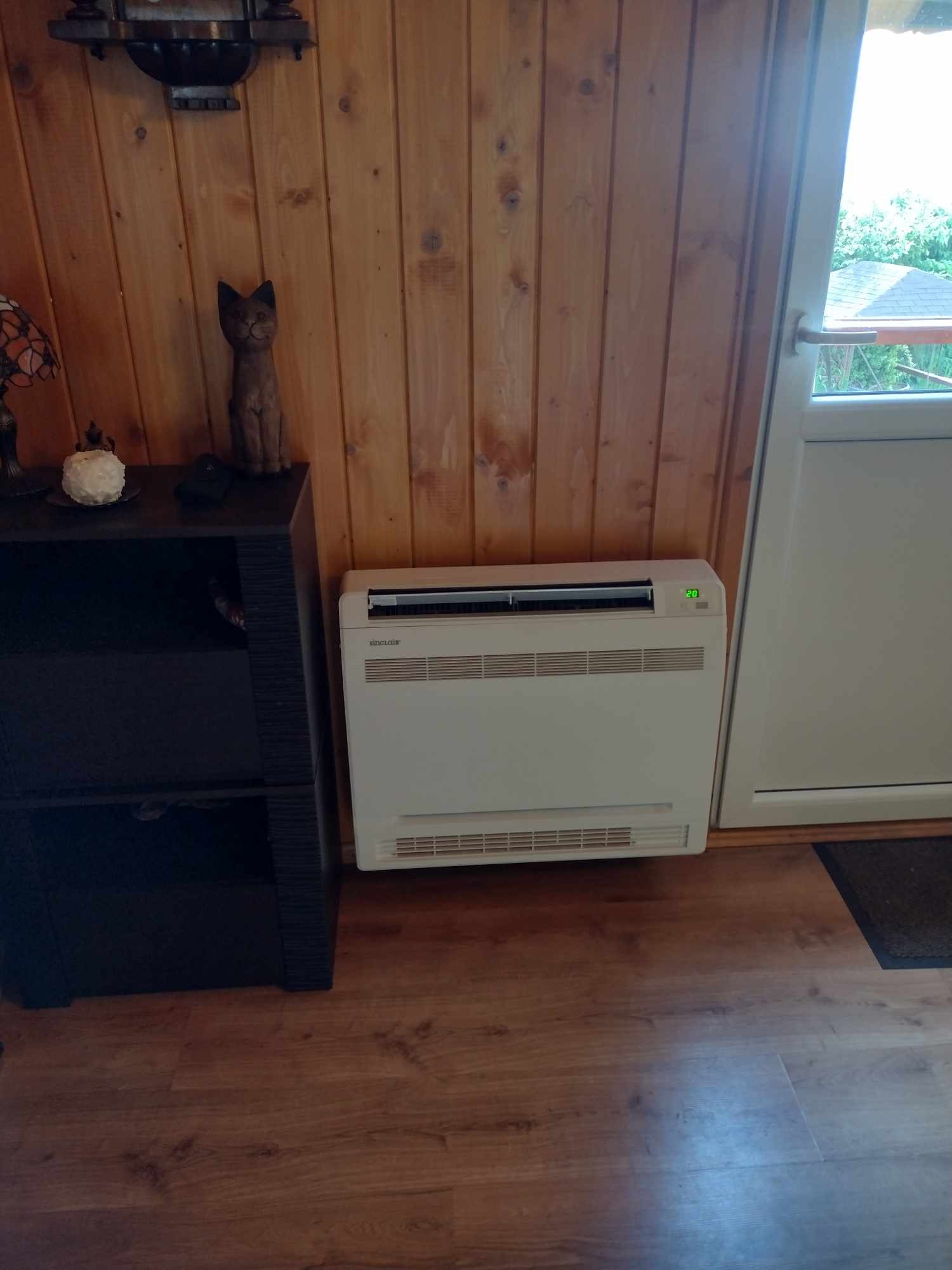 Podłogowy klimatyzator Sinclair z cyfrowym wyświetlaczem temperatury, umieszczony przy drewnianej ścianie i drzwiach wejściowych w pomieszczeniu z drewnianą podłogą.