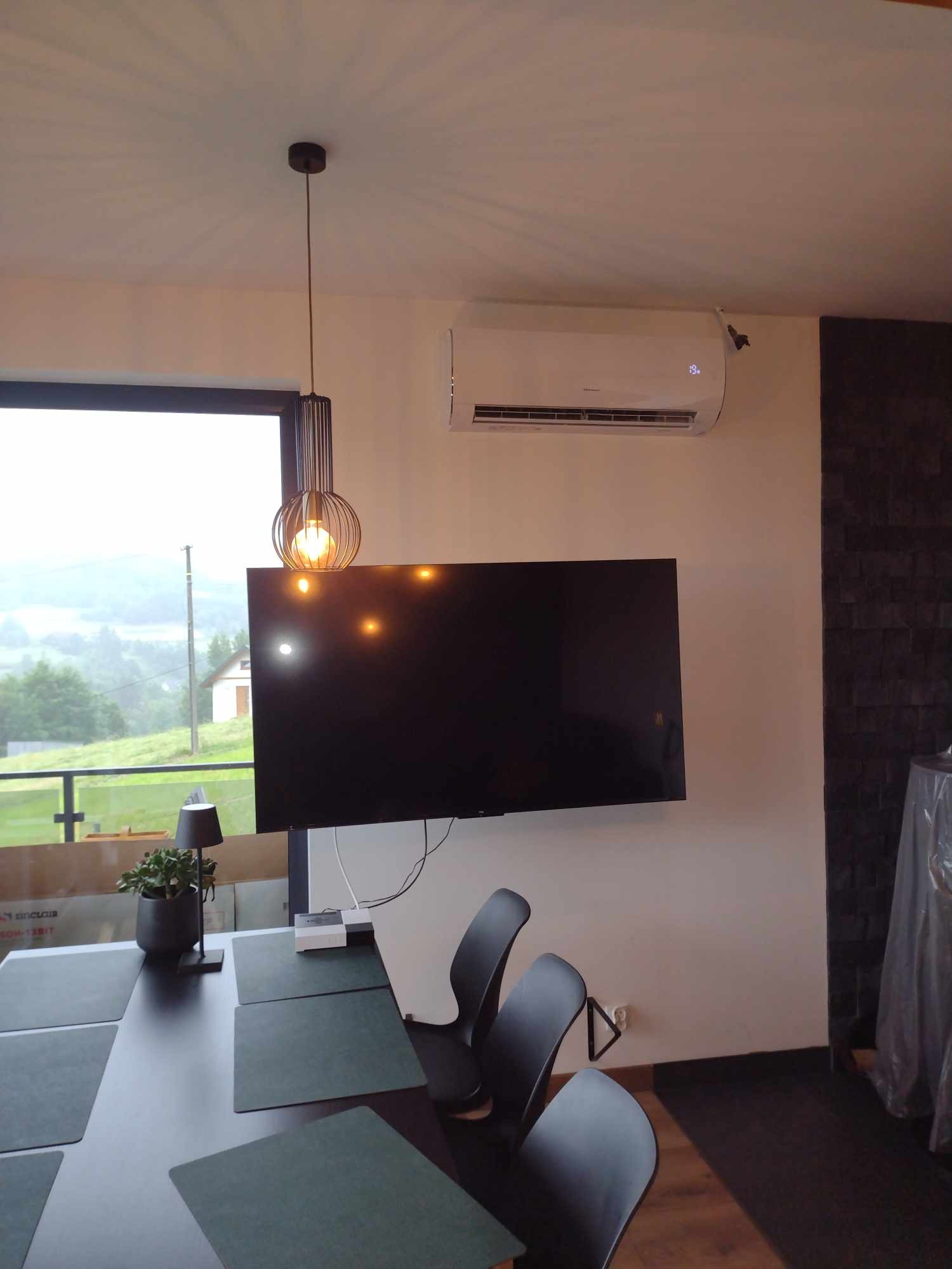 Nowoczesne wnętrze jadalni z zamontowaną klimatyzacją ścienną nad telewizorem i widokiem na zielone wzgórza za oknem.