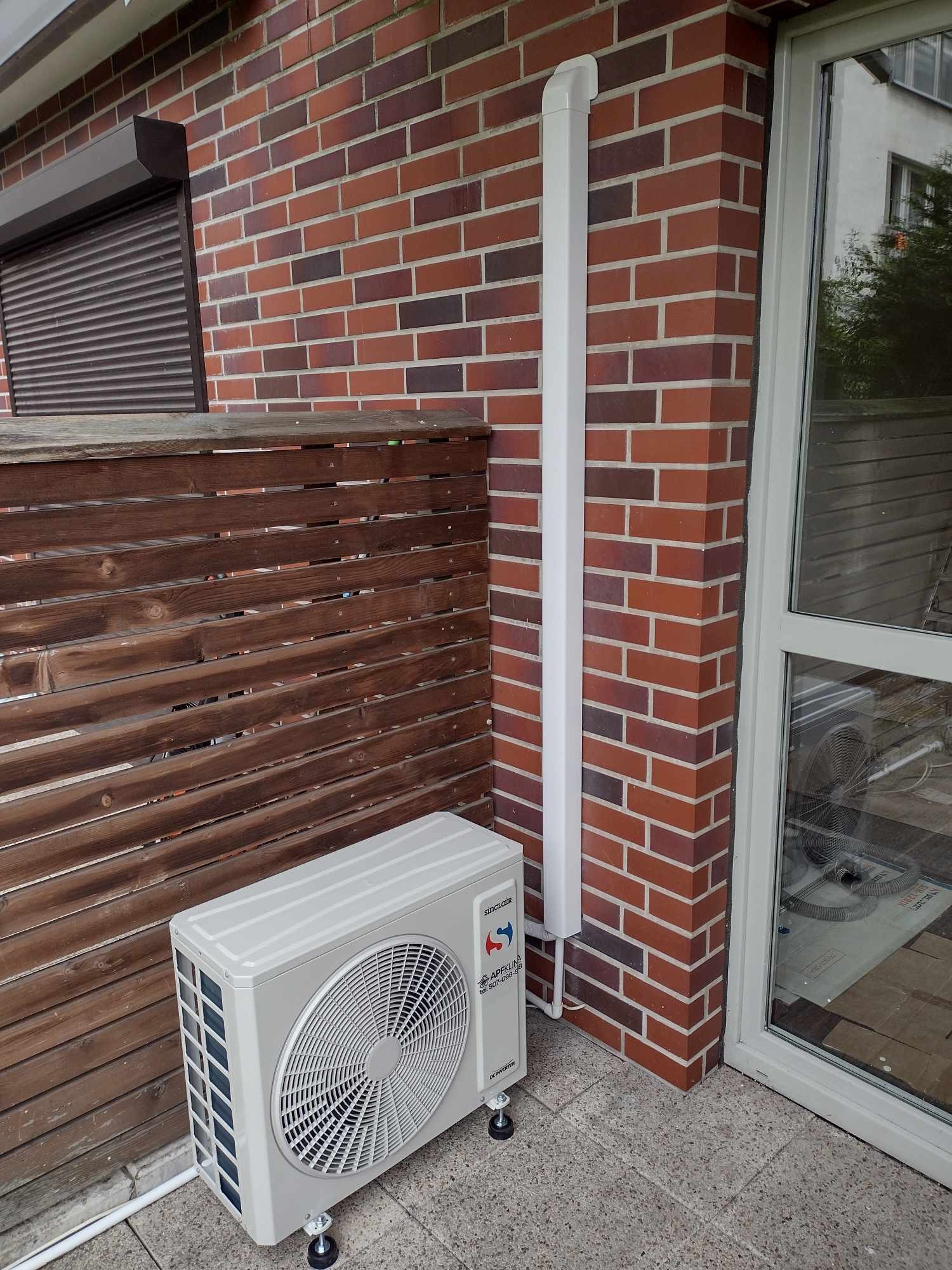Zewnętrzna jednostka klimatyzacji Sinclair zamontowana na zewnątrz budynku z czerwoną cegłą, widoczna biała rura odprowadzająca skropliny, brązowe żaluzje w oknie, całość na szarym brukowanym podłożu.
