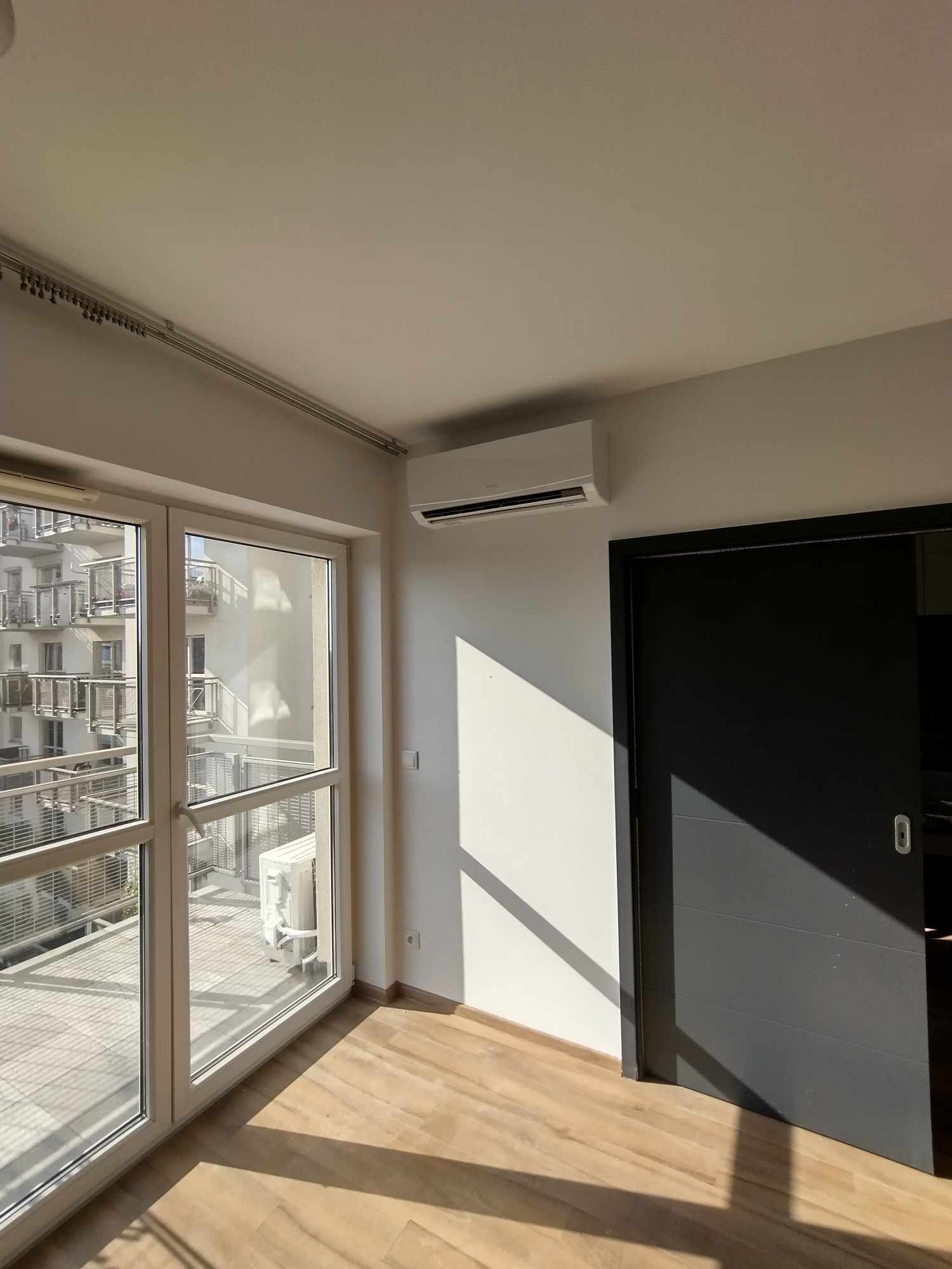 Wnętrze jasnego pokoju z zamontowanym klimatyzatorem ściennym, widok na balkon z apartamentowcem w tle przez duże okno, promienie słoneczne padają na podłogę i ścianę.