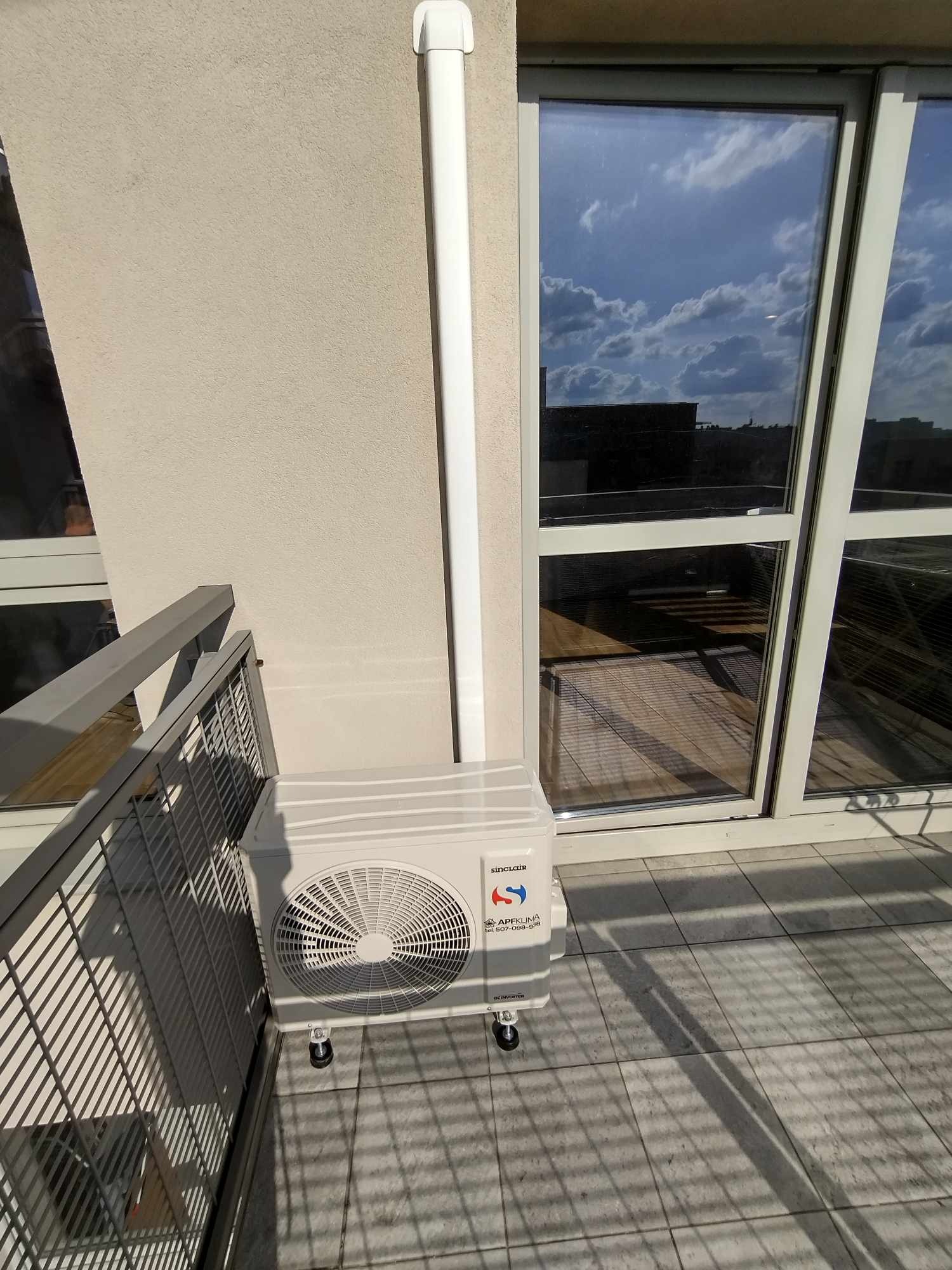 Jednostka zewnętrzna klimatyzatora Sinclair na kółkach, ustawiona na balkonie, widoczna instalacja odprowadzająca skropliny, w tle okno balkonowe z odbijającym się niebem.