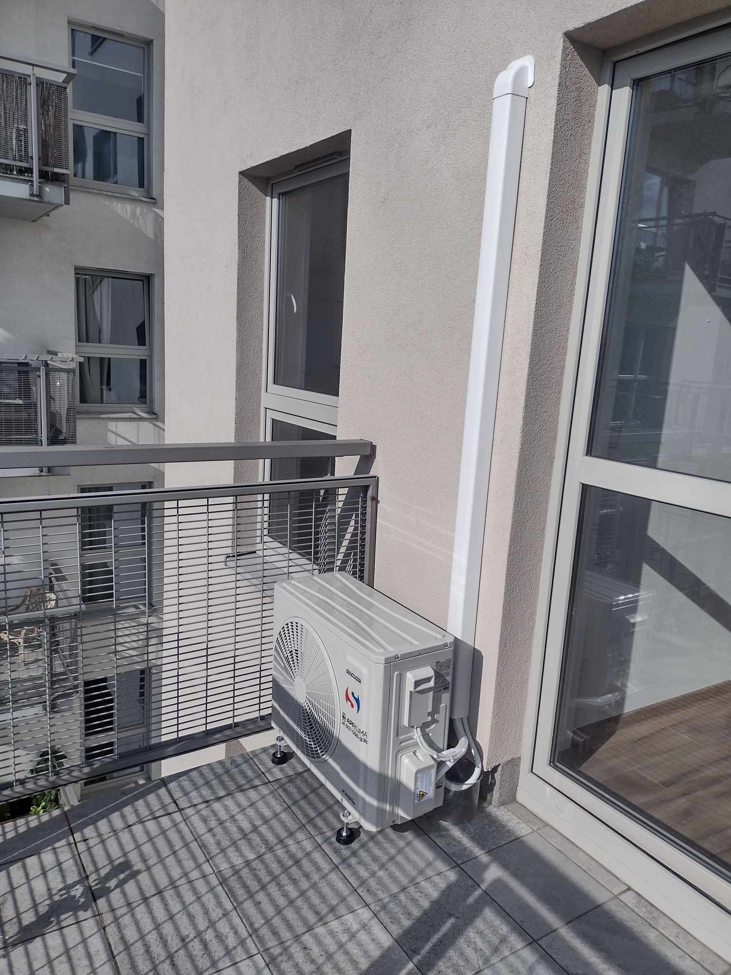 Jednostka zewnętrzna klimatyzatora Sinclair zamontowana na balkonie, z widocznymi przewodami i rurą odprowadzającą skropliny, na tle jasnej elewacji budynku i balustrady balkonu.