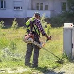 Dan-instal - Mężczyzna w odblaskowej kamizelce i czapce przycina trawę kosiarką spalinową na nierównym terenie przed budynkiem mieszkalnym. Widoczny biały pojemnik na kosiarkę.