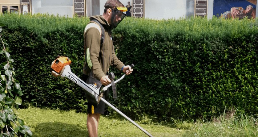 Mężczyzna w masce ochronnej przycina żywopłot za pomocą podkaszarki spalinowej Stihl, w tle zielony trawnik i budynek z muralem.