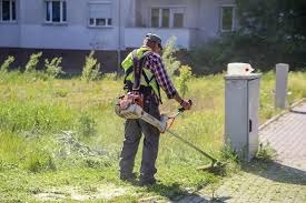 Mężczyzna w odblaskowej kamizelce i czapce przycina trawę kosiarką spalinową na nierównym terenie przed budynkiem mieszkalnym. Widoczny biały pojemnik na kosiarkę.
