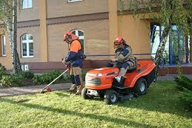 Dwóch mężczyzn w kaskach ochronnych i odzieży roboczej pielęgnuje trawnik przed budynkiem: jeden kosi trawę traktorkiem, drugi podcina krawędzie podkaszarką.