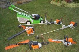 Zestaw sprzętu ogrodniczego na trawie: kosiarka z zielonym korpusem, dwie podkaszarki spalinowe z białymi uchwytami i pomarańczowymi elementami, pilarka spalinowa z pomarańczową obudową oraz nożyce...